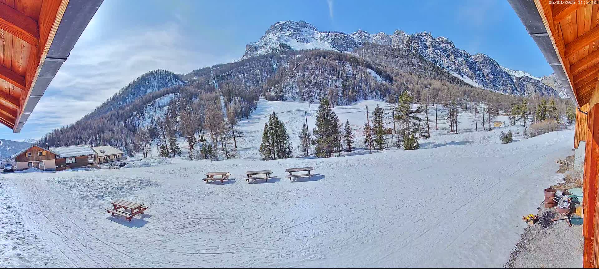 A snow-covered landscape with several picnic tables, a few buildings, and a mountain range in the background under a mostly sunny sky.