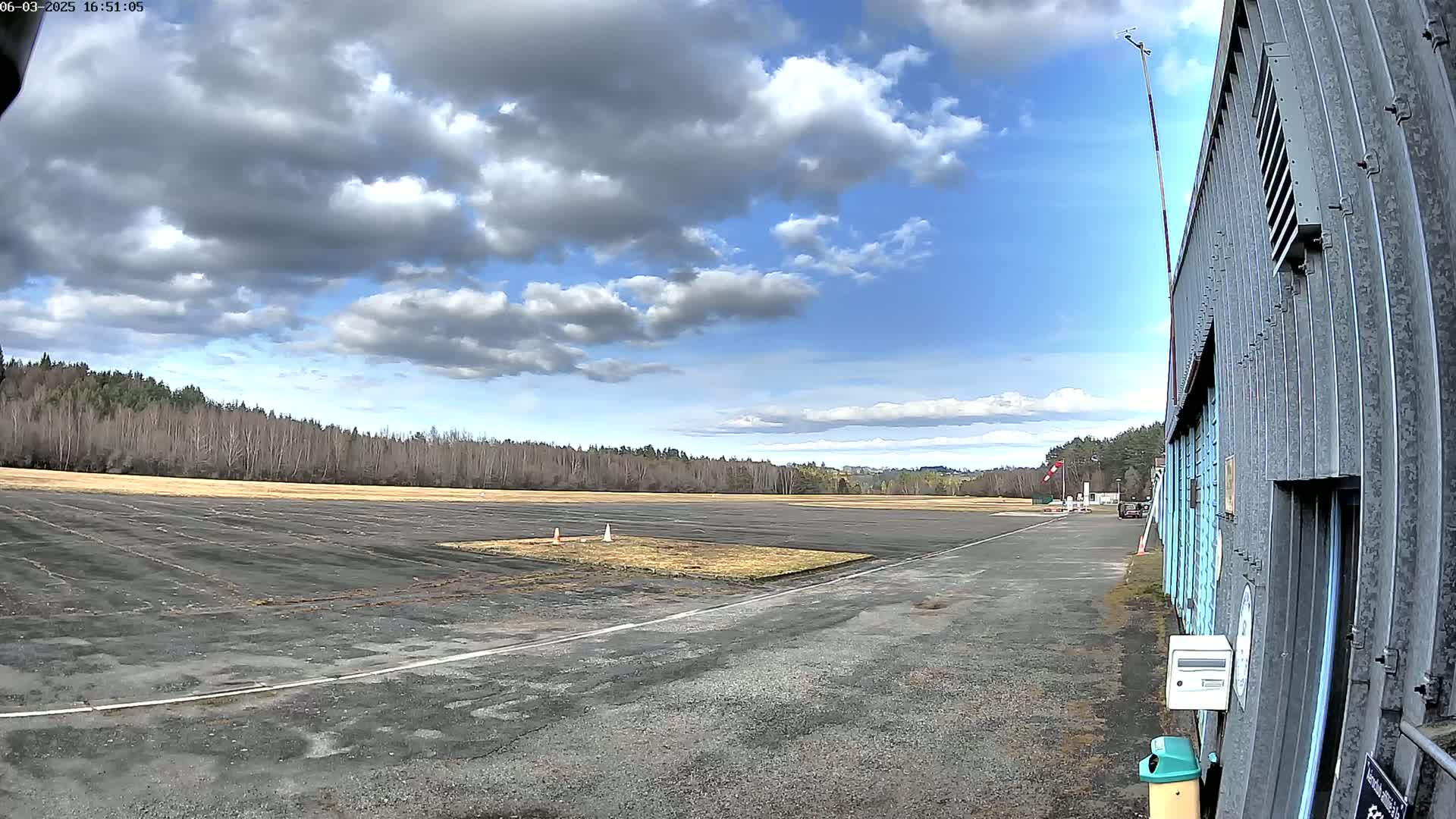 A partly cloudy sky overlooks a paved airfield next to a hangar and a line of trees.