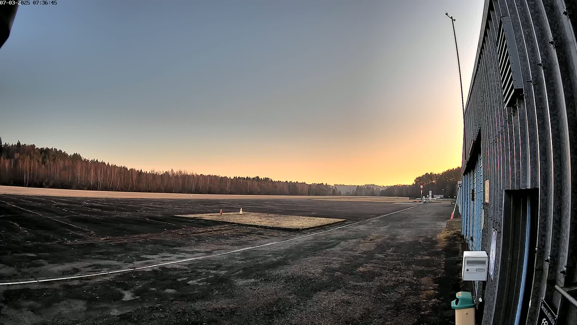 A dark asphalt airfield at sunrise, with a line of bare trees in the background under a clear, pale orange and blue sky.