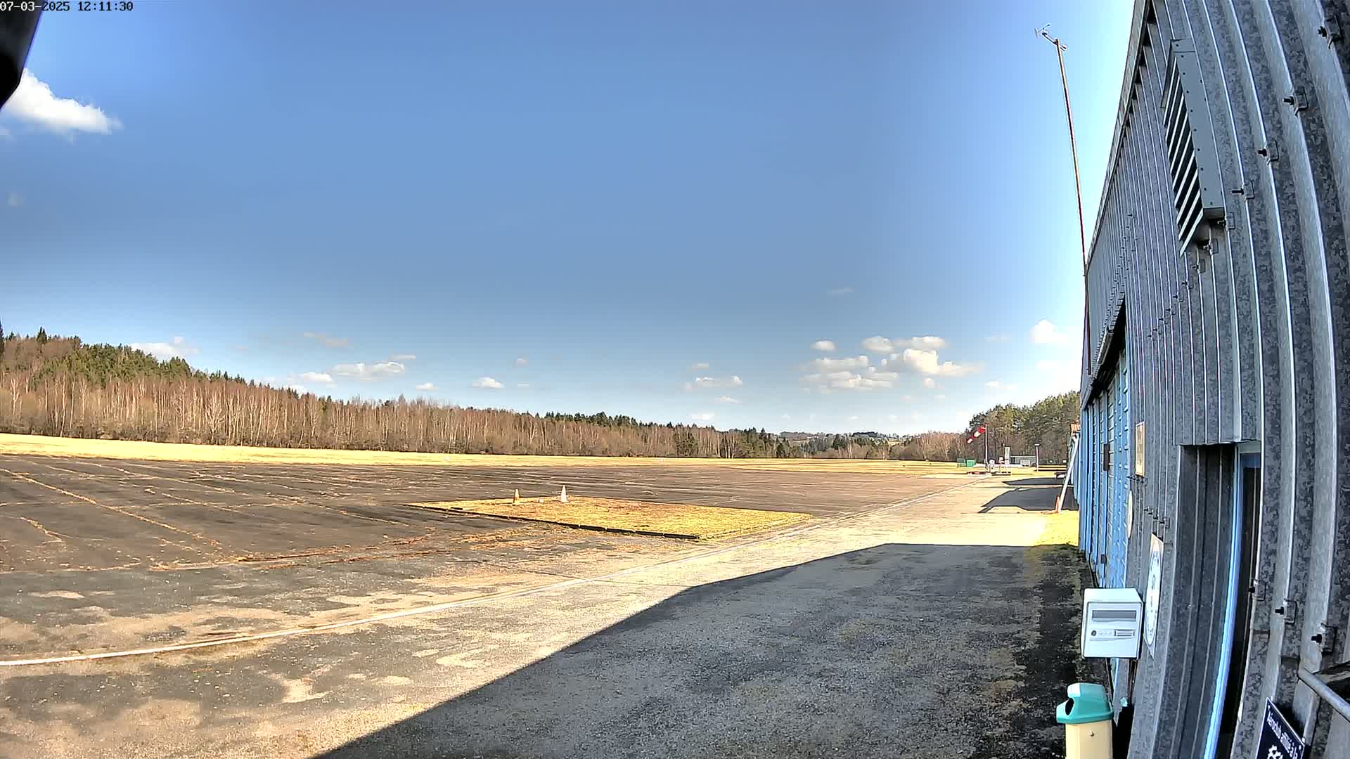 A mostly sunny day shows a paved airfield bordered by trees, with a metal building on the right.