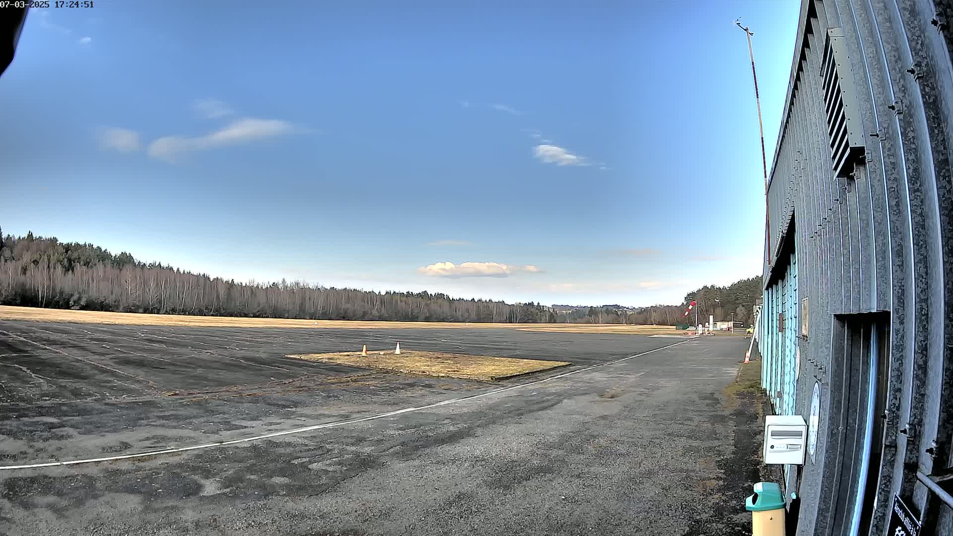 A mostly clear blue sky overlooks a small airfield with a paved runway and a hangar, bordered by a line of leafless trees.