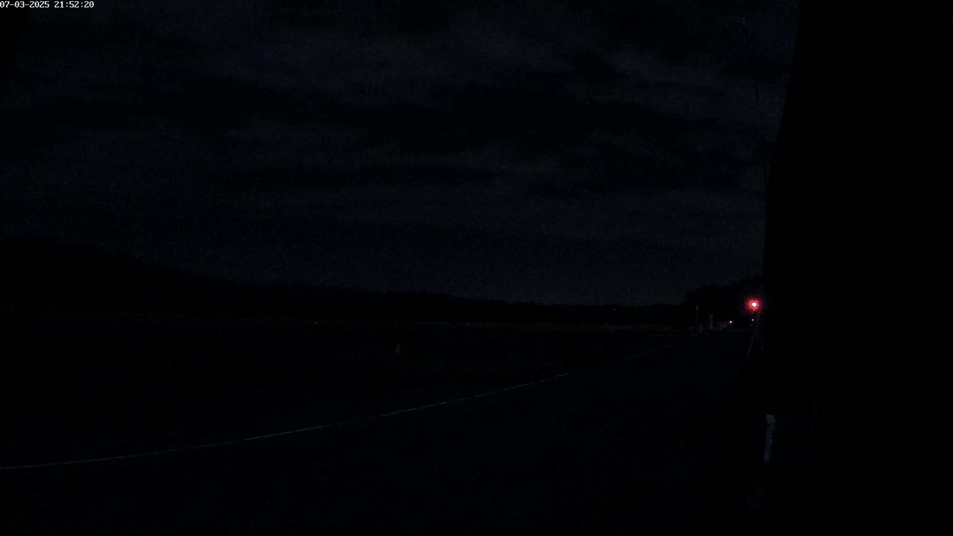 A dark, nighttime scene shows a flat, dark road stretching into the distance under a cloudy sky, with a single faint red light visible in the far distance.