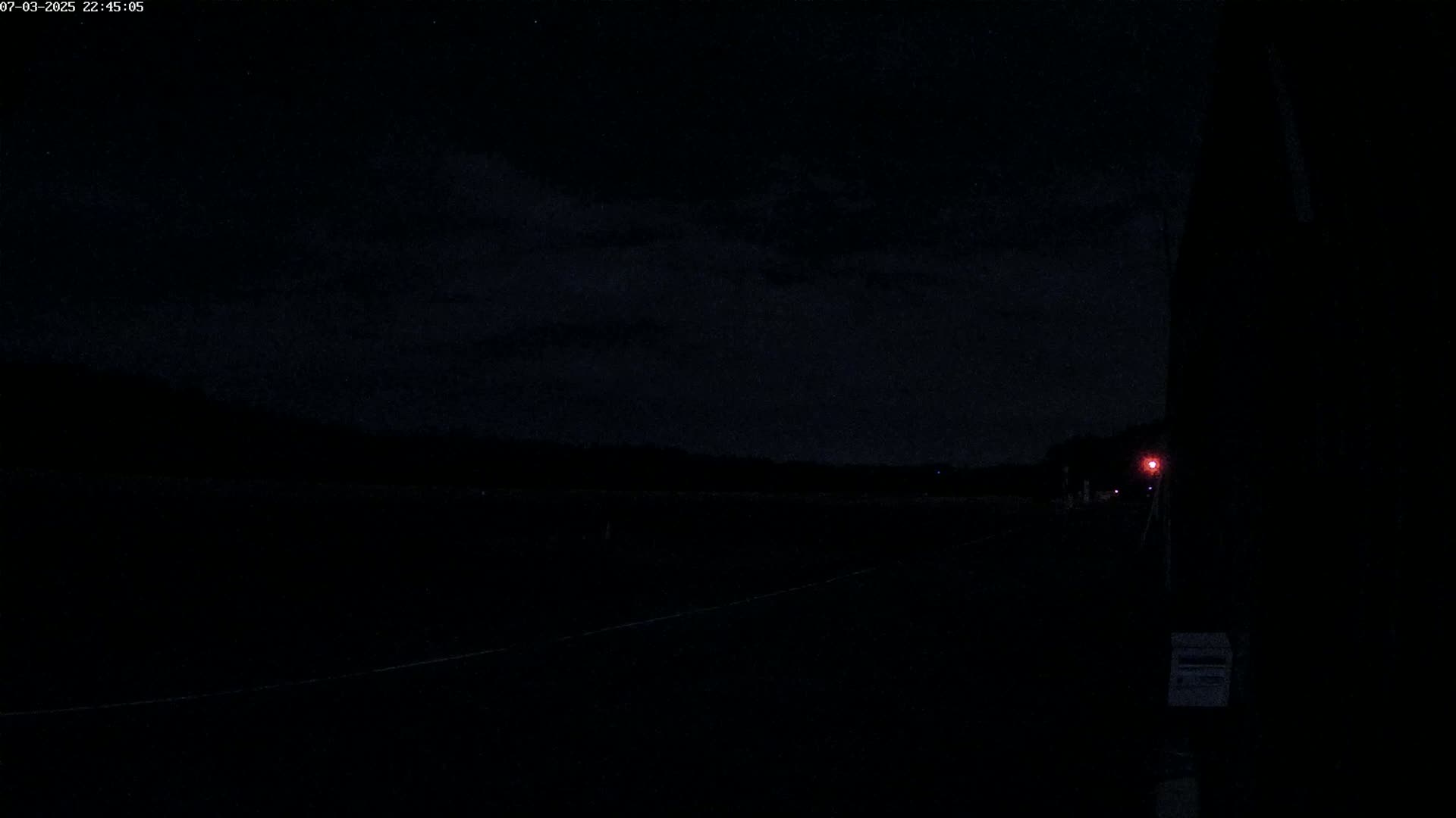 A dark, nighttime scene shows a flat, empty expanse of land under a cloudy sky with a single faint red light visible in the distance.