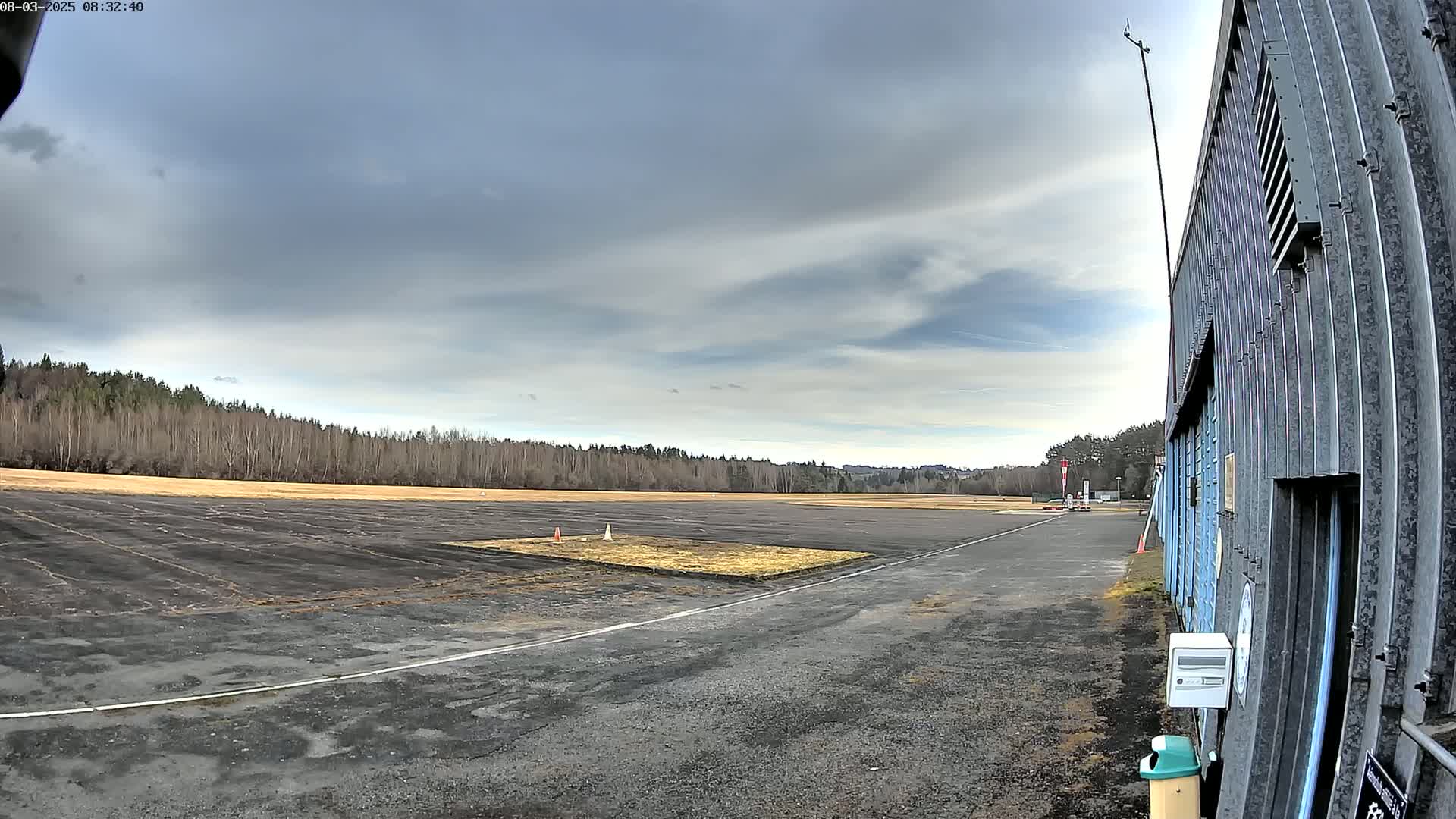 A paved airfield with a grassy patch in the center extends to a line of trees under a mostly cloudy sky.