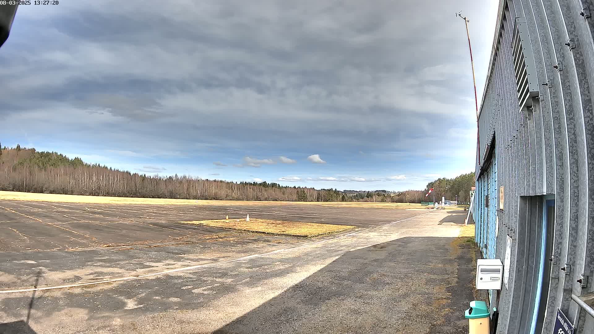 A mostly cloudy day reveals a tarmac airfield bordered by a line of trees, with a hangar visible to the right.