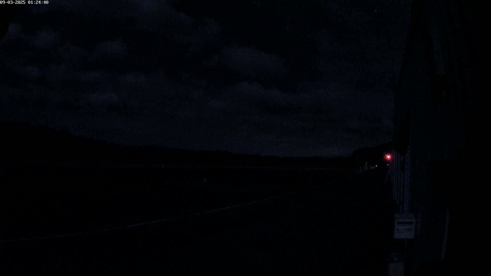 A dark, nighttime scene shows a paved road stretching into the distance under a cloudy sky, with a single distant red light visible.