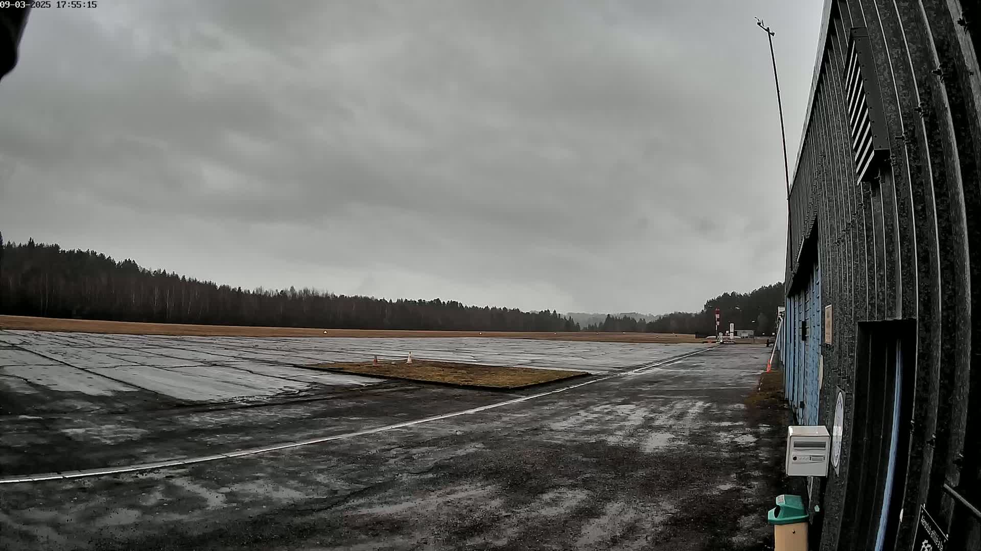 A wet, paved airfield stretches out under a cloudy sky, bordered by a line of trees and a hangar to the right.