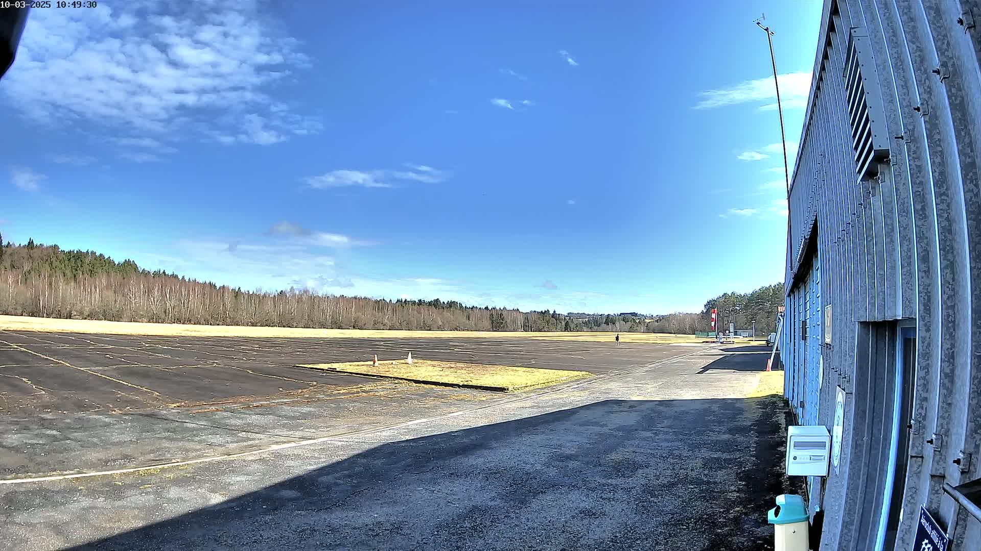 A mostly sunny day reveals a paved airfield bordered by trees, with a single person visible in the distance and a corrugated metal building at the lower right.
