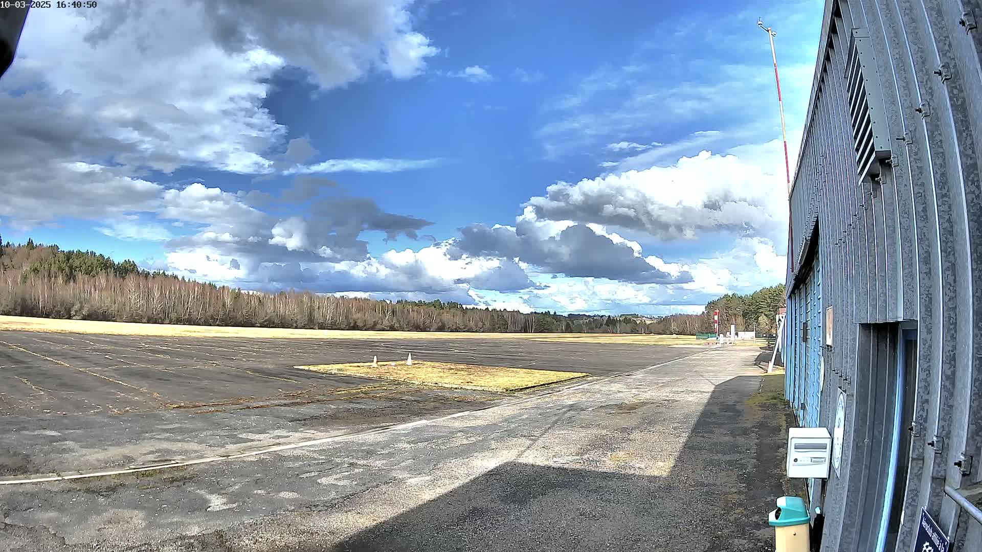 A mostly sunny day with puffy white clouds shows a view of a small airfield with a hangar in the foreground.