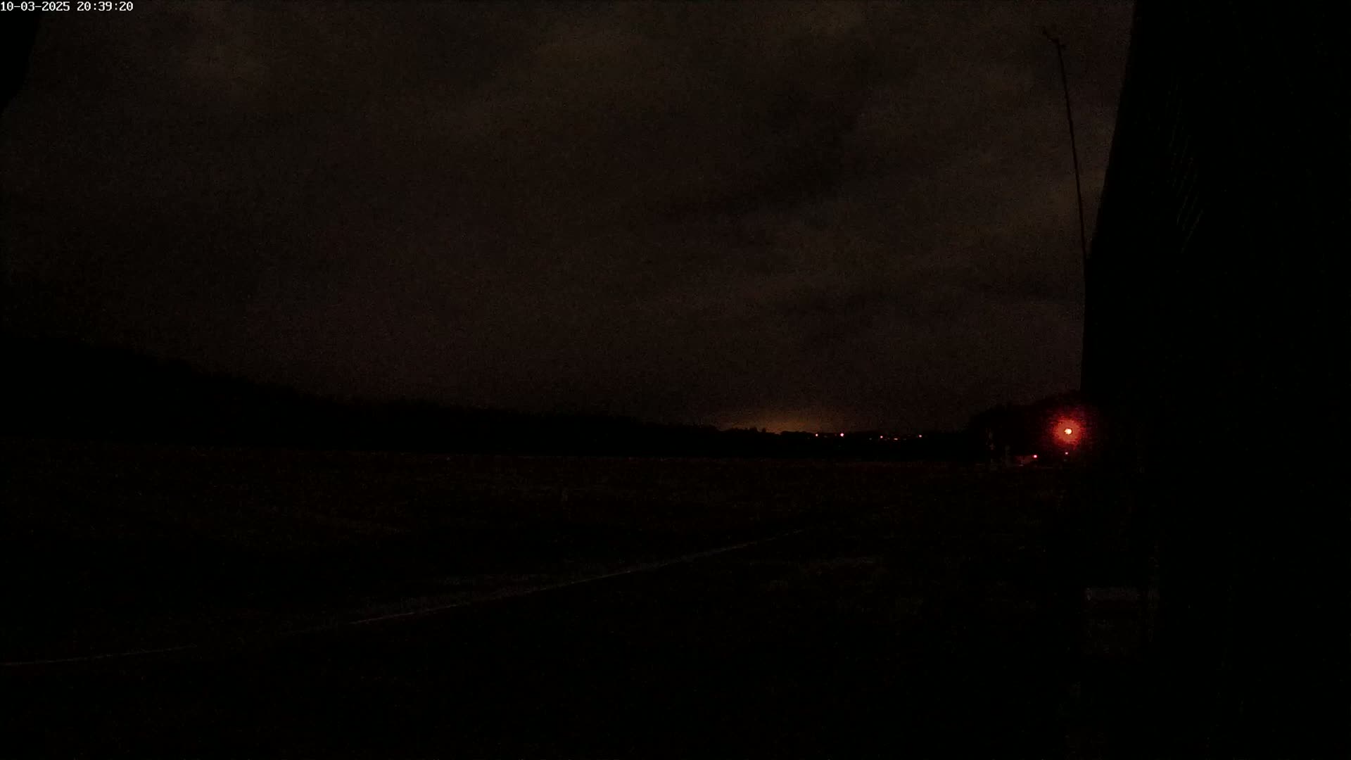 A dark, nighttime scene shows a flat, dark landscape under a mostly cloudy sky, with a few faint lights visible in the distance and a single red light closer to the viewer.