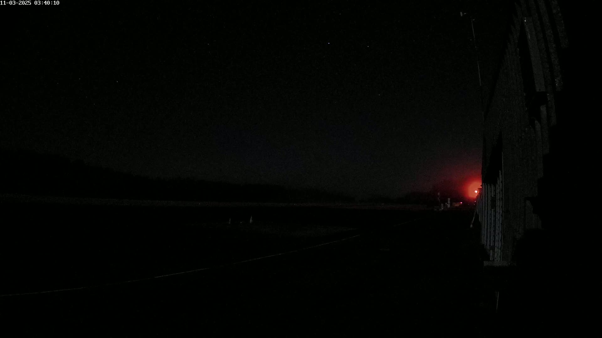 A dark, nighttime scene shows a flat, empty field next to a tall, dark structure, with a faint red light visible in the distance under a clear, starry sky.