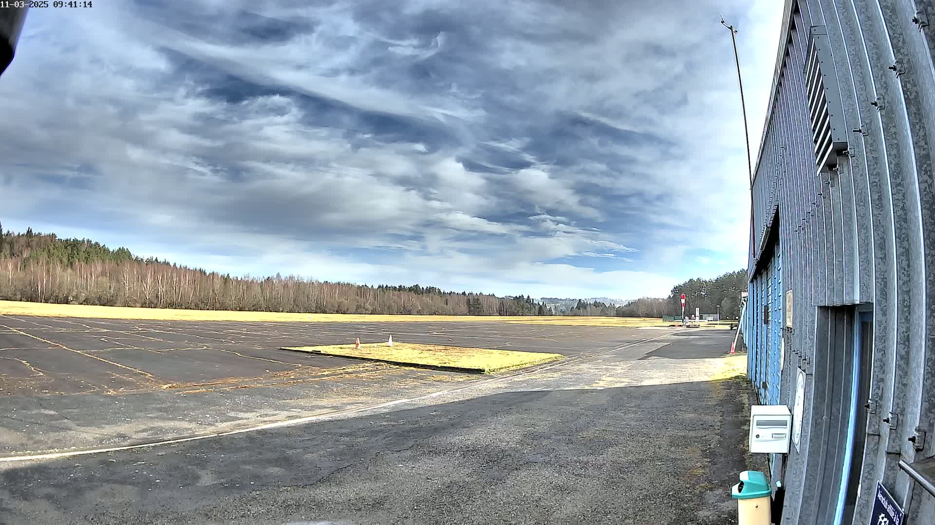 A mostly clear sky with scattered clouds overlooks a small airfield with a large hangar to the right, and a line of trees in the background.