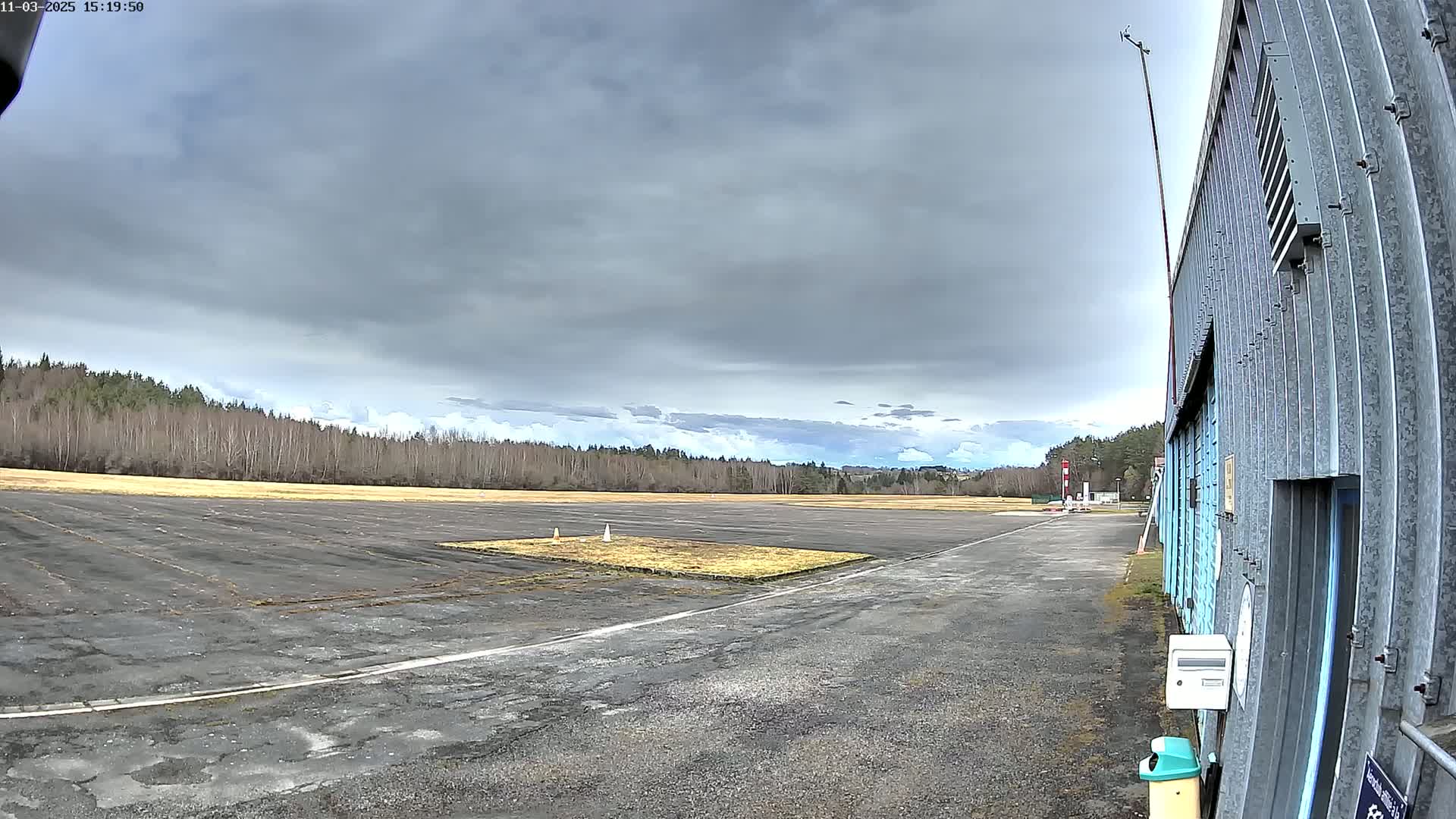 A paved airfield, bordered by a line of leafless trees under a cloudy sky, stretches toward a metal hangar in the foreground.