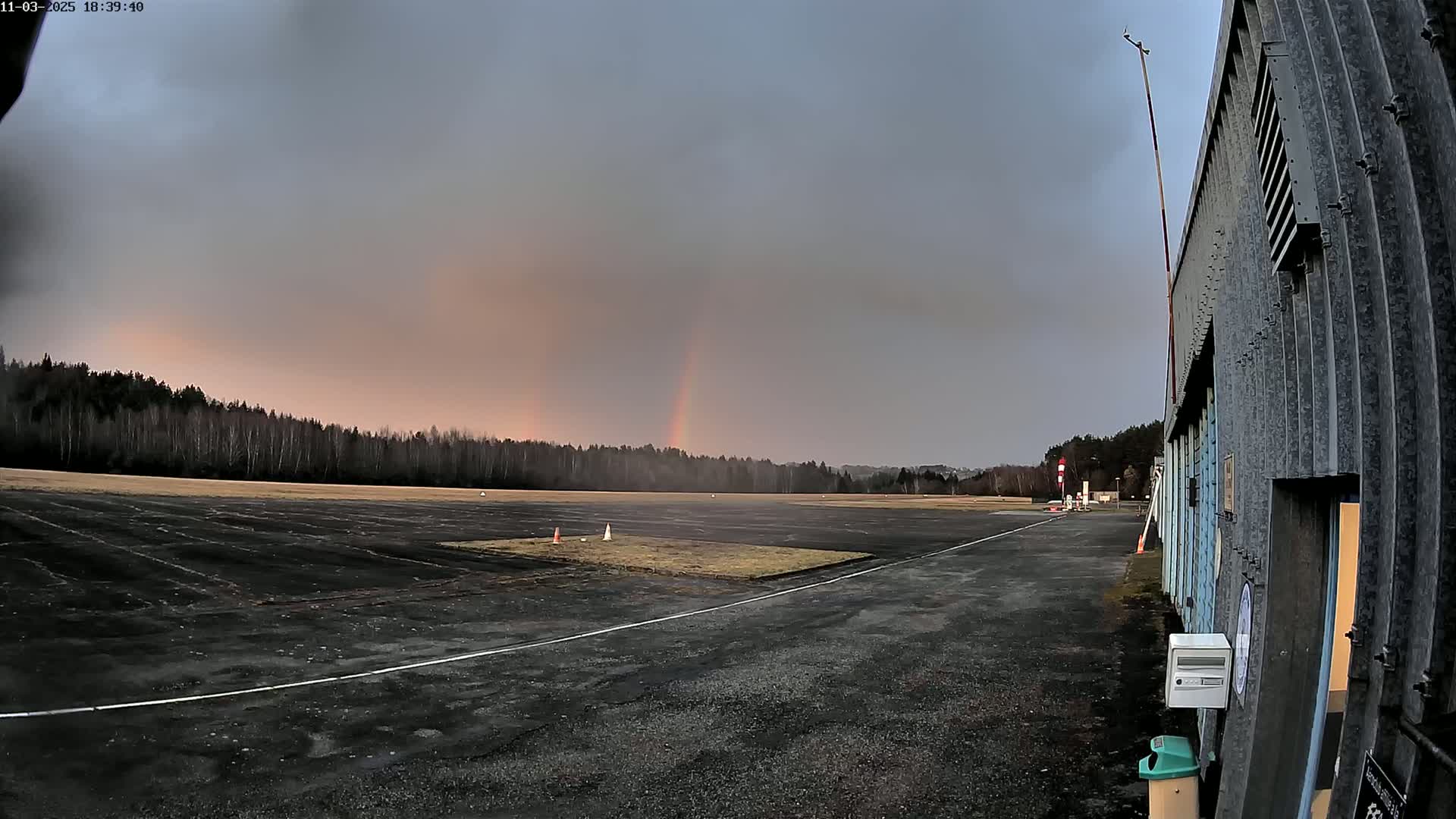 A faint rainbow arcs over a dark tarmac airfield bordered by a line of trees under a mostly cloudy sky.