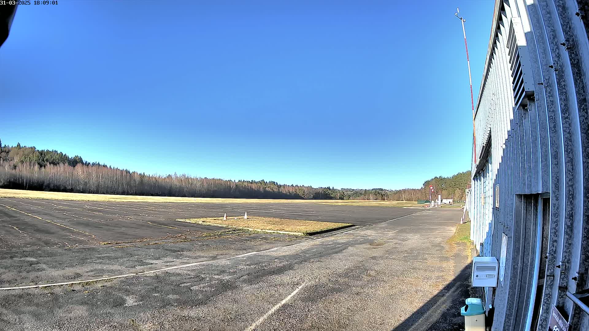 A mostly empty airfield with a dark asphalt surface and a line of trees in the background under a clear blue sky.