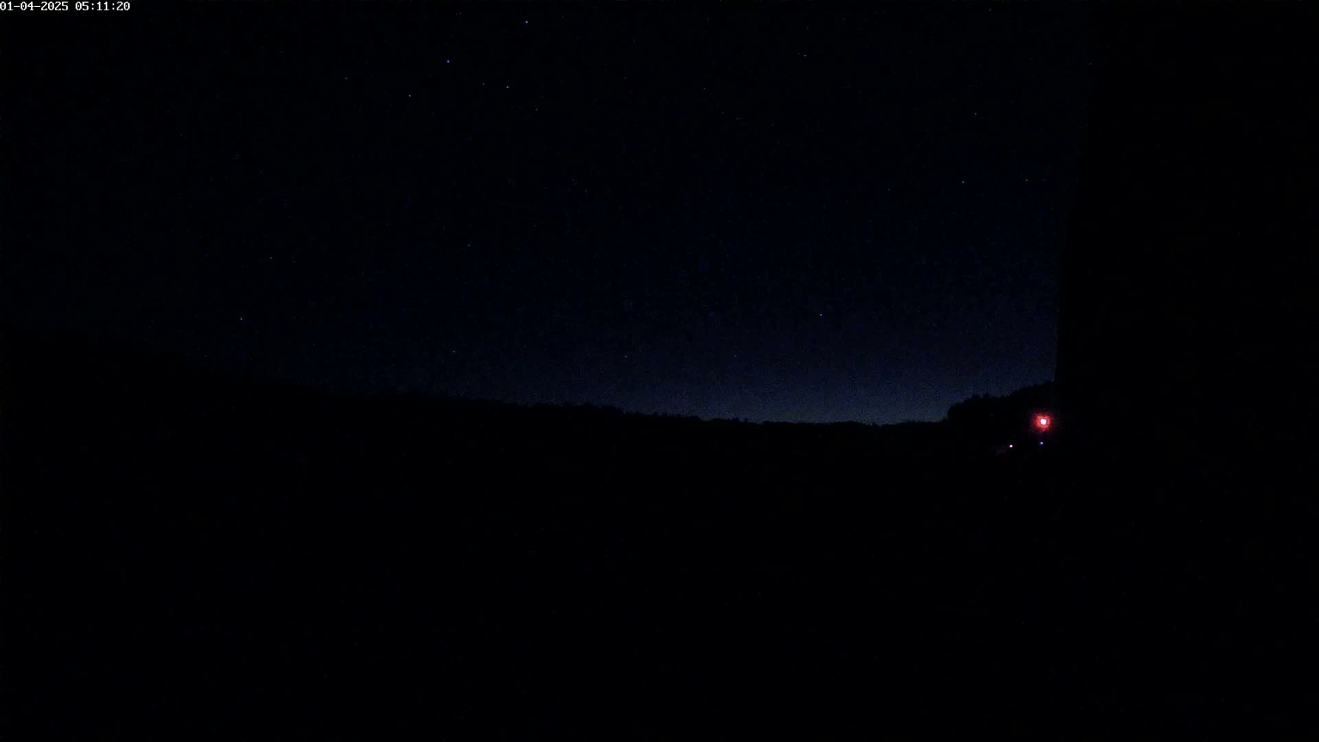 A dark, starlit night sky above a silhouetted landscape, with a single faint red light visible in the lower right corner.