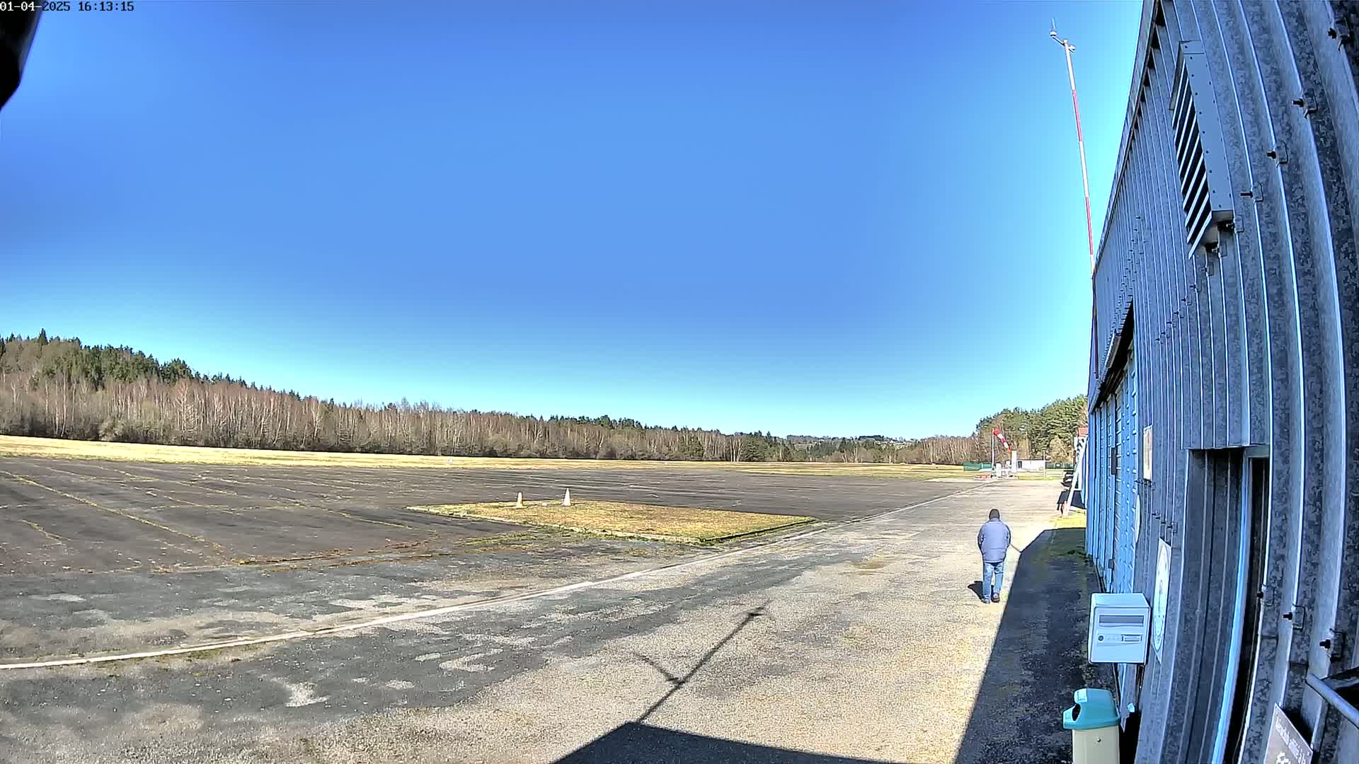 A person walks past a hangar toward a large, empty tarmac area bordered by a line of trees under a clear blue sky.