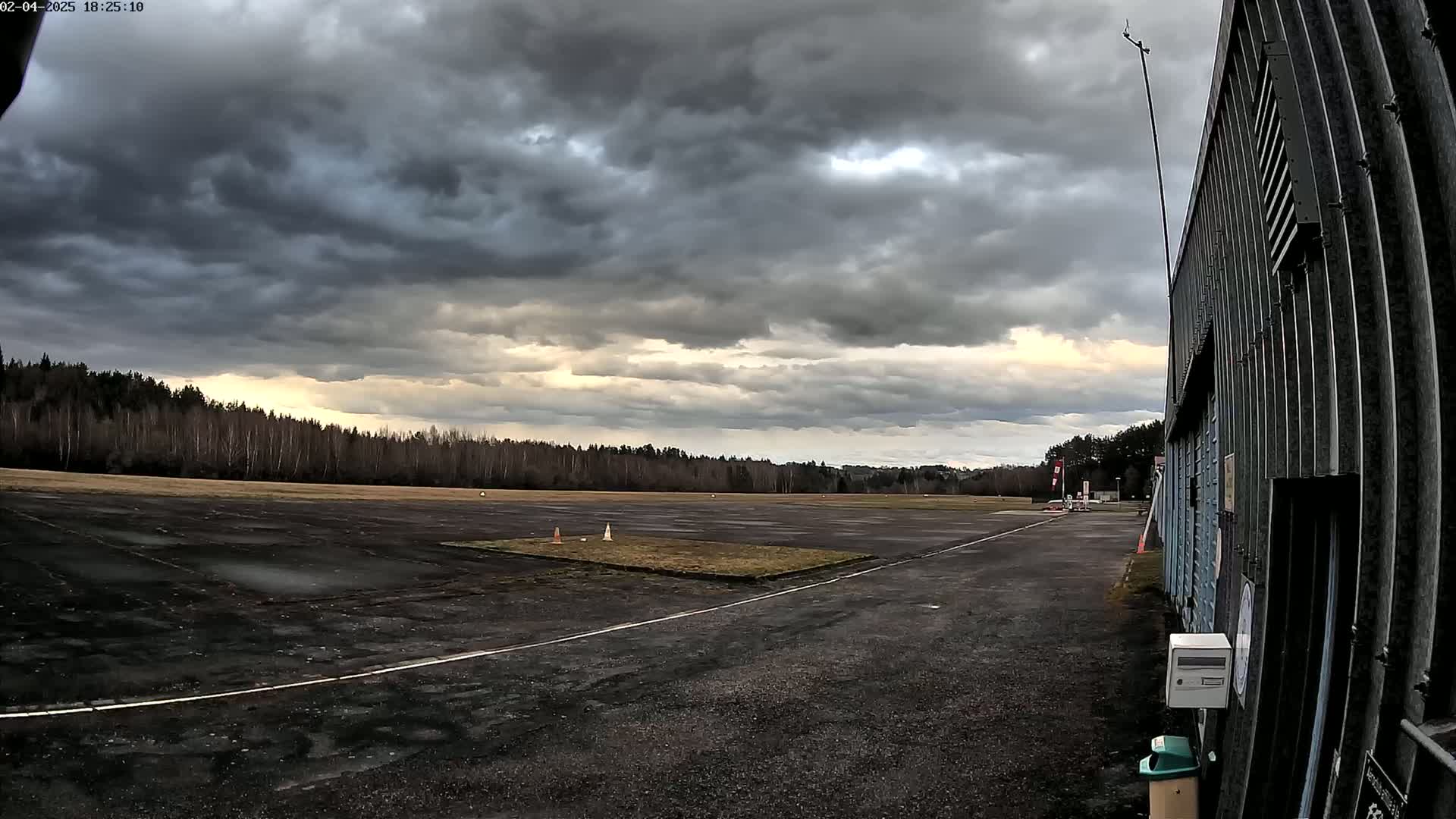 A dark, overcast sky hangs over a deserted tarmac area next to a tall corrugated metal building, bordered by a line of leafless trees.