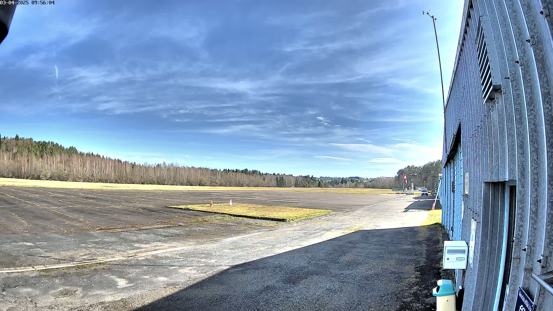 A mostly sunny day shows a paved airfield bordered by a line of trees, next to a corrugated metal building.