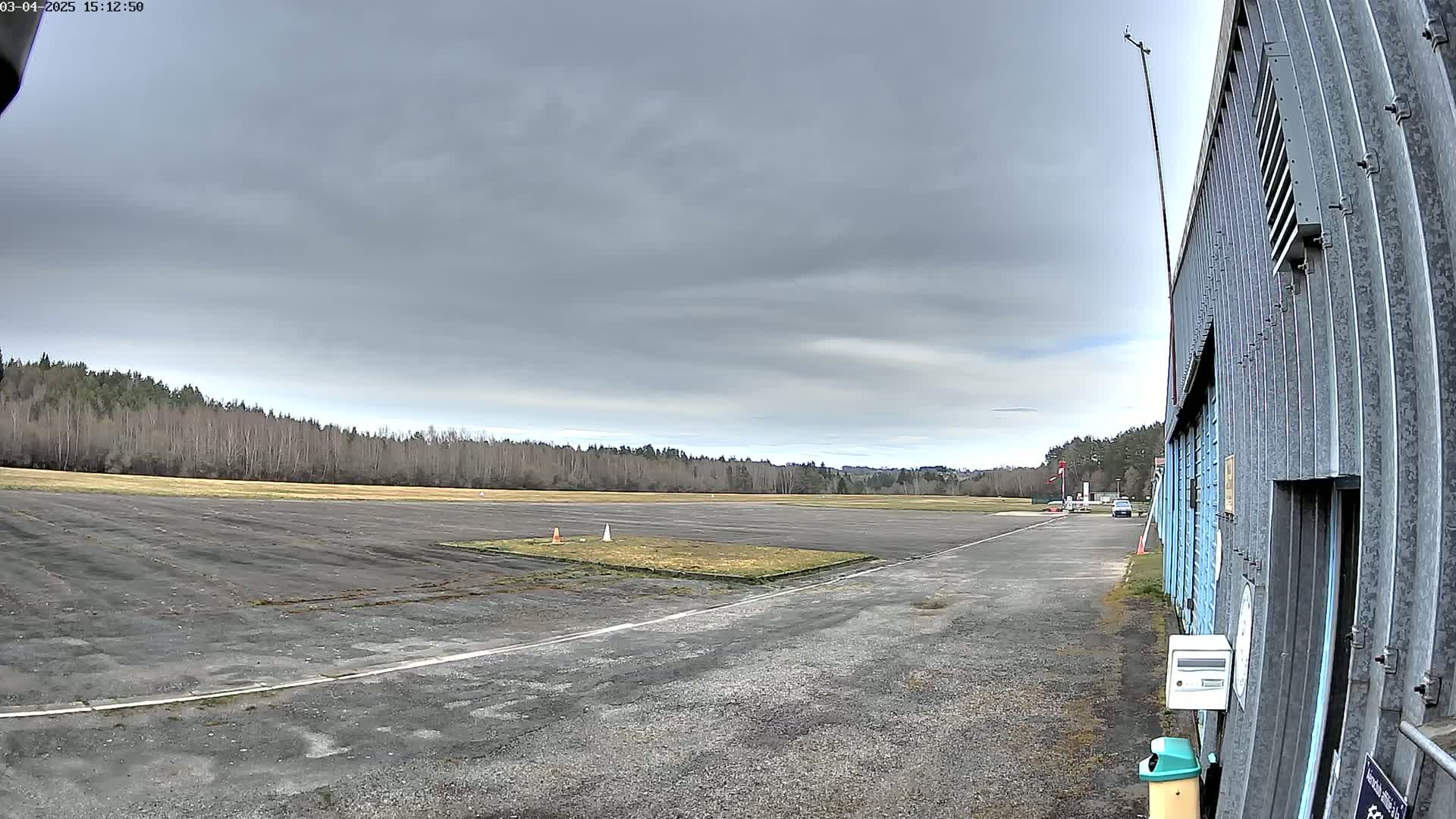 A mostly overcast sky hangs over a small airfield with a paved runway and a line of trees in the background, next to a corrugated metal hangar.