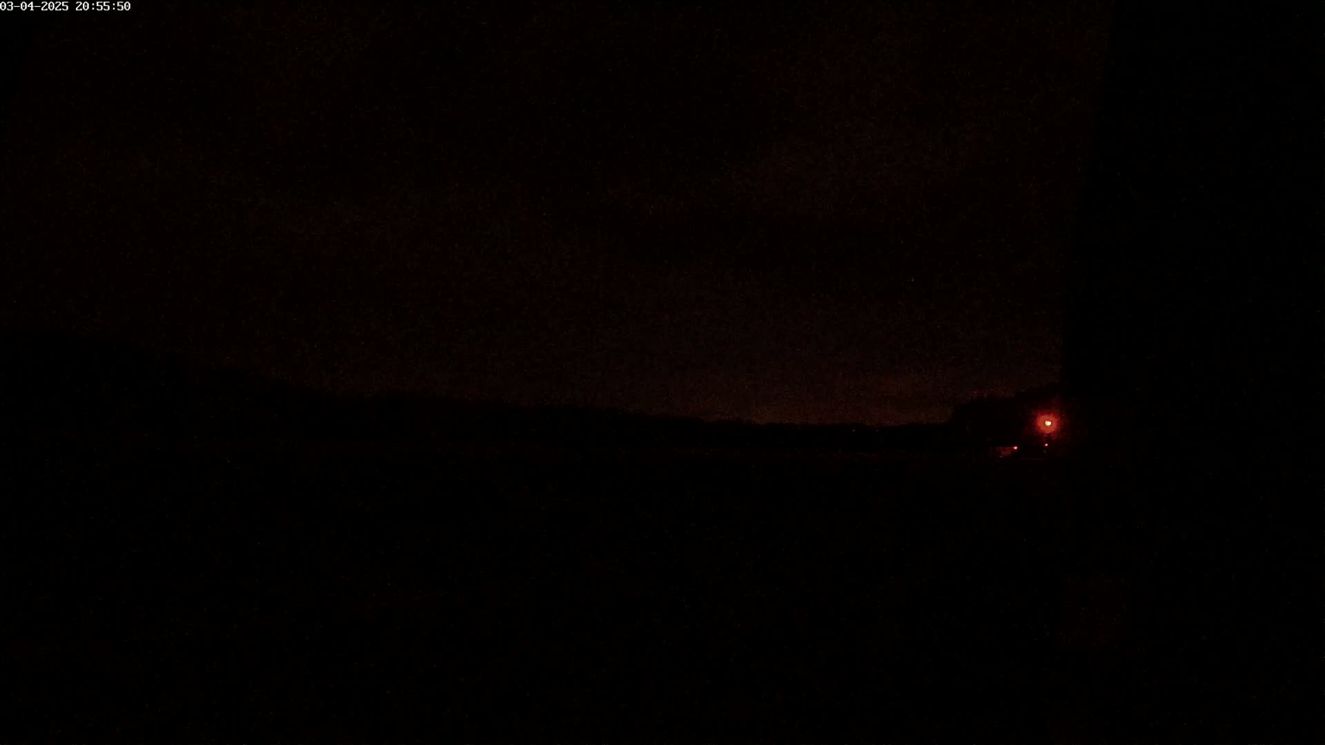 A dark nighttime scene shows a faint horizon line and a single reddish light in the distance under a mostly clear sky.