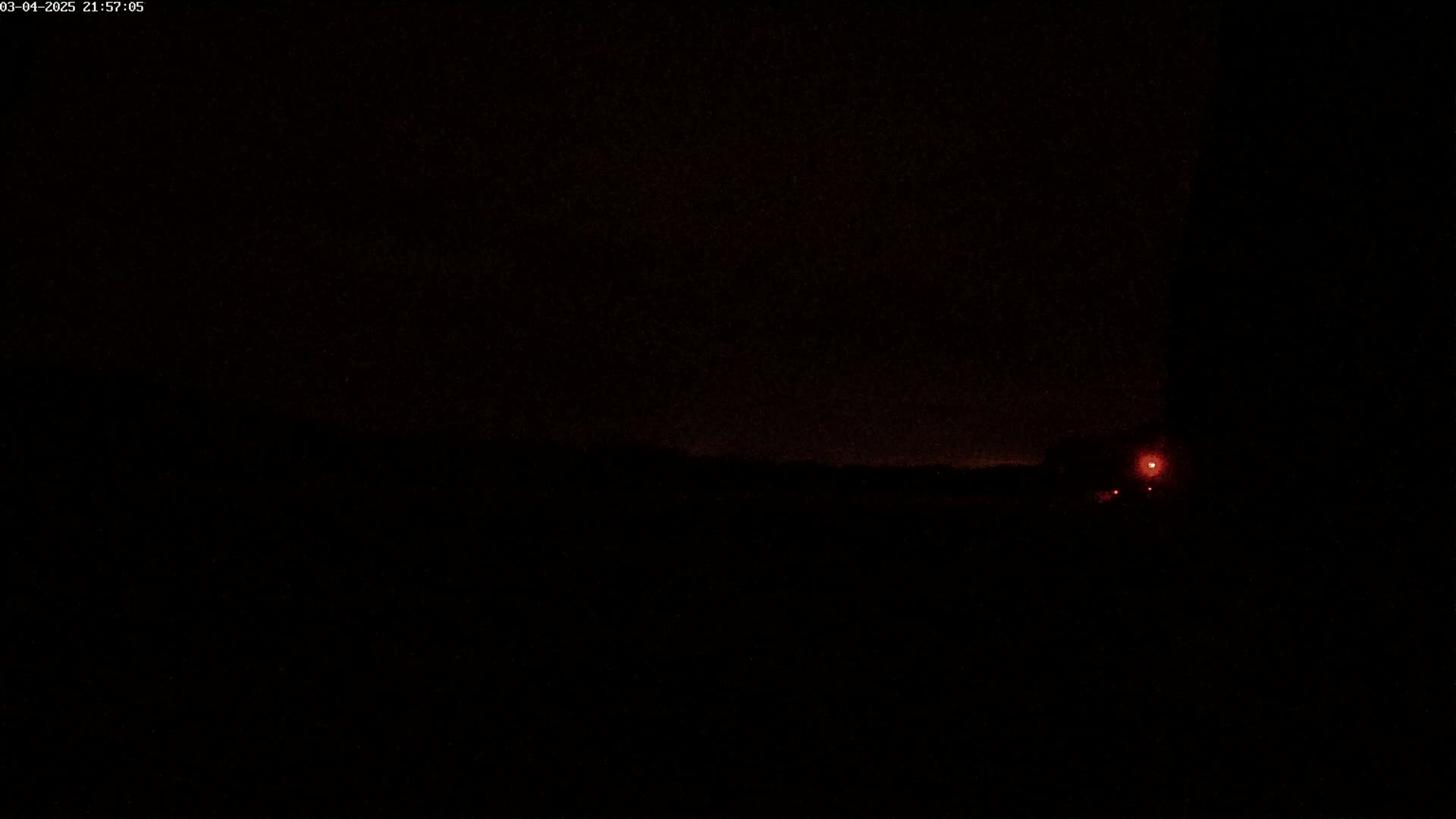 A nighttime outdoor scene shows a dark landscape with faint red lights visible in the distance under a mostly clear sky.