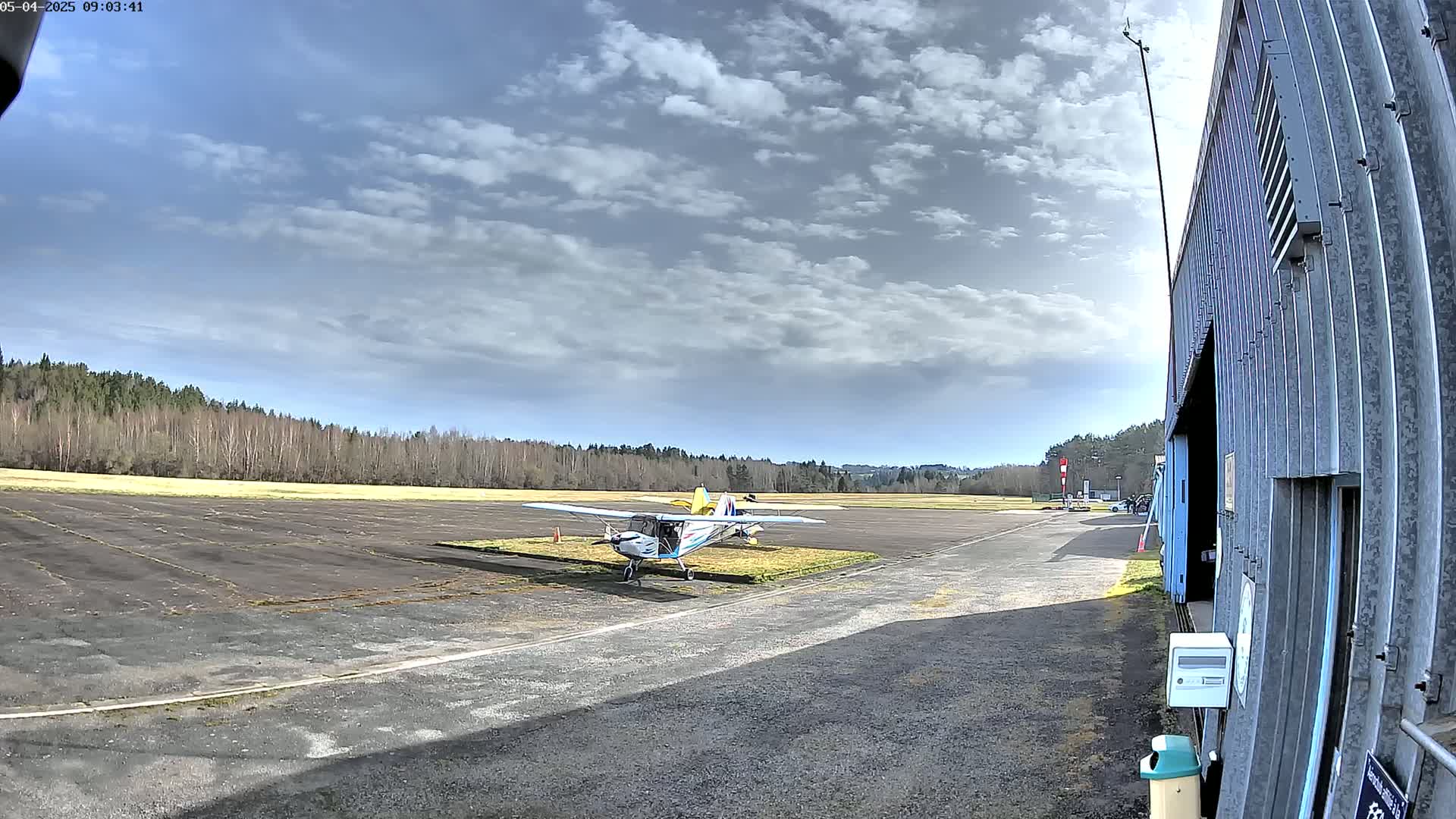A small airplane sits on a tarmac next to a hangar, under a partly cloudy sky.