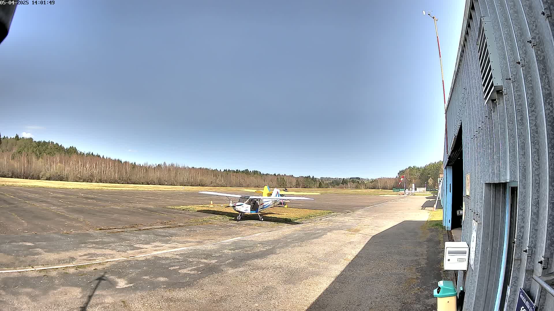 A small airplane sits on a tarmac next to a hangar, under a clear blue sky in front of a line of trees.