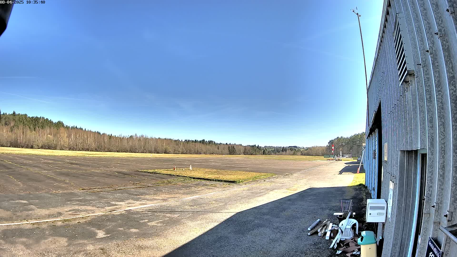 A paved airfield stretches out under a clear blue sky, bordered by a line of trees and a hangar to the right.