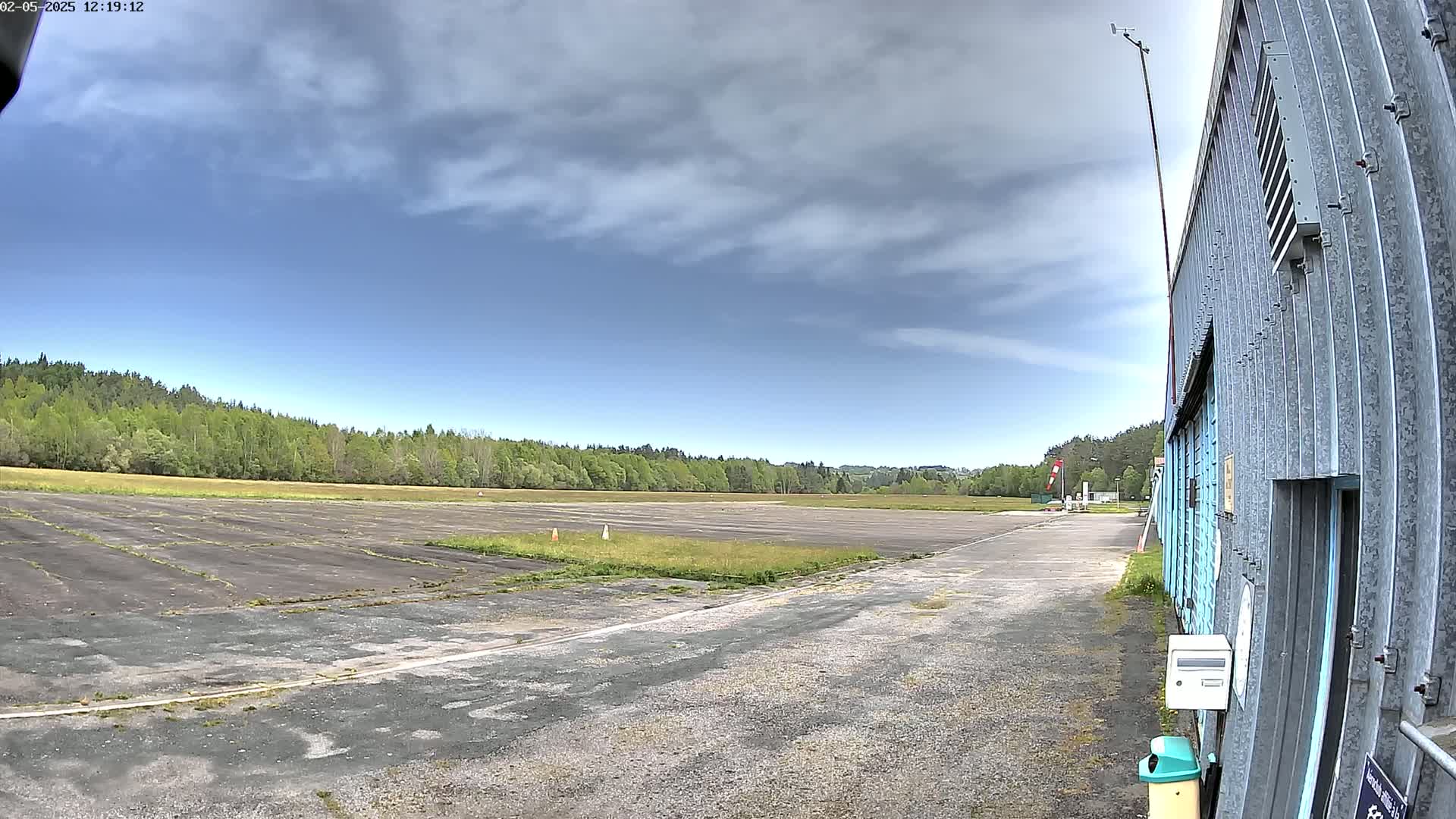 A partly cloudy sky overlooks a paved airstrip, bordered by trees and a hangar.