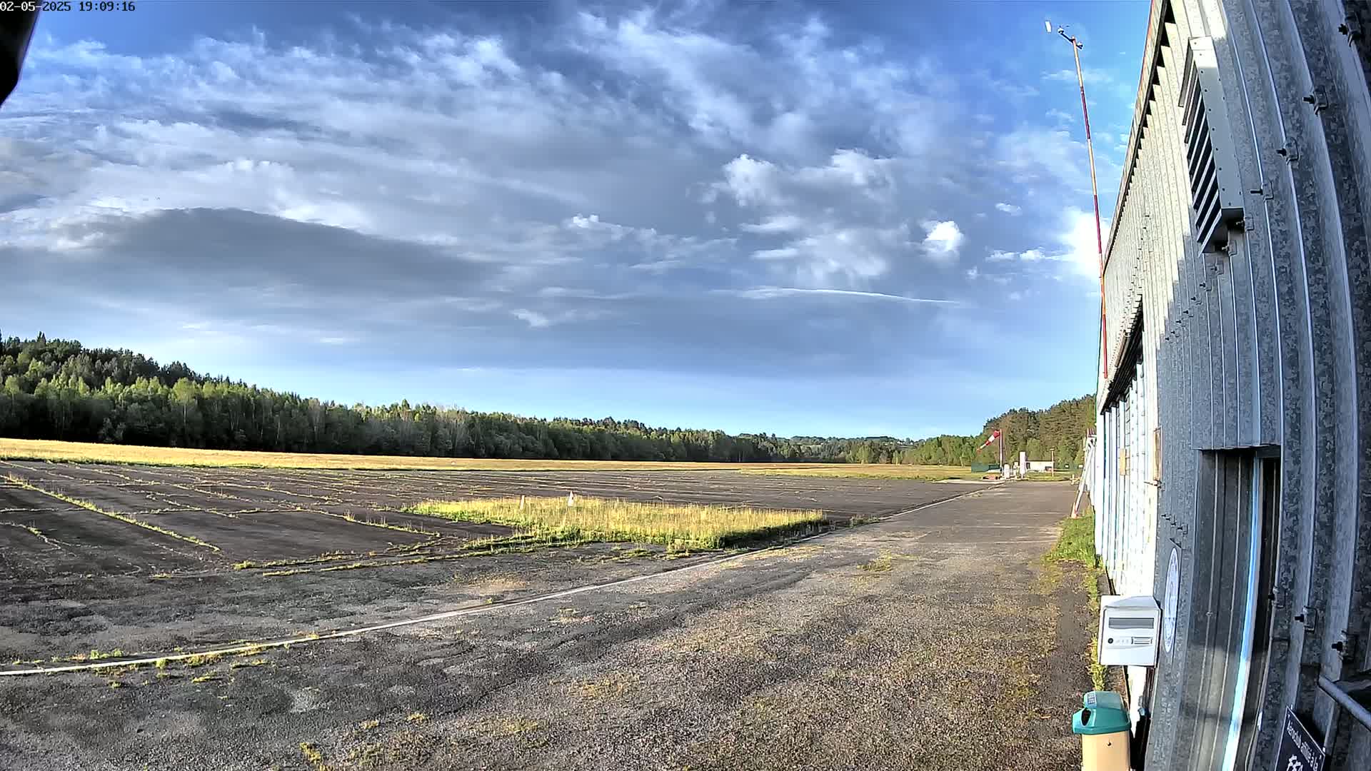 A mostly sunny day reveals a paved runway next to a hangar, with a grassy field and a line of trees in the background.