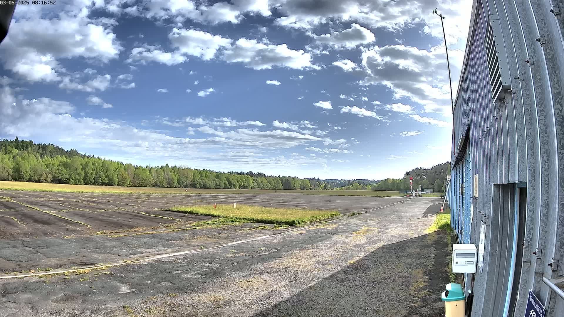 A partly sunny day reveals a paved airfield bordered by a line of trees, with a large metal building to the right.