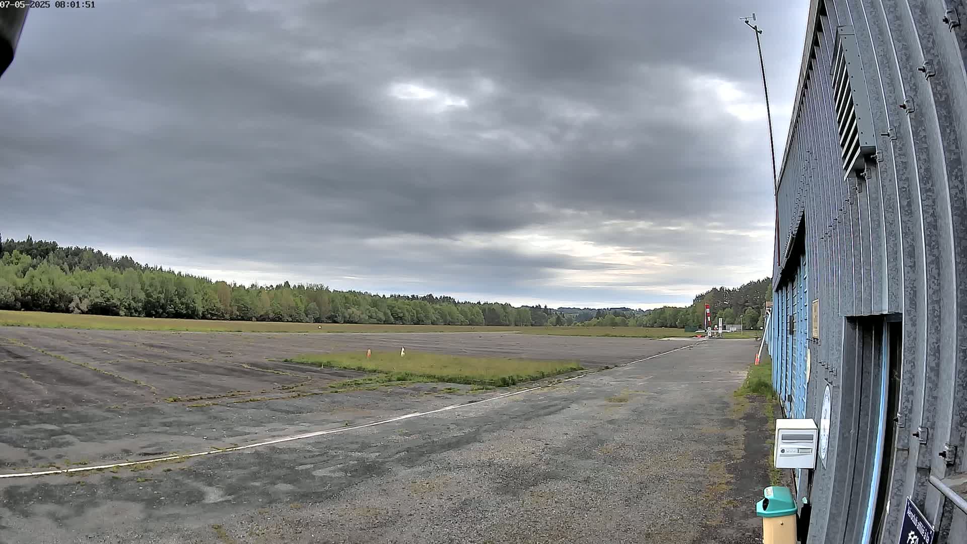 A mostly overcast sky hangs over a small airfield with a long, empty tarmac runway and a line of trees in the background.