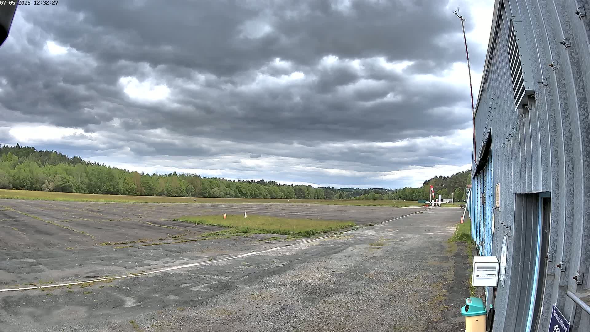 A mostly overcast sky hangs over a small, grassy airfield next to a corrugated metal building.