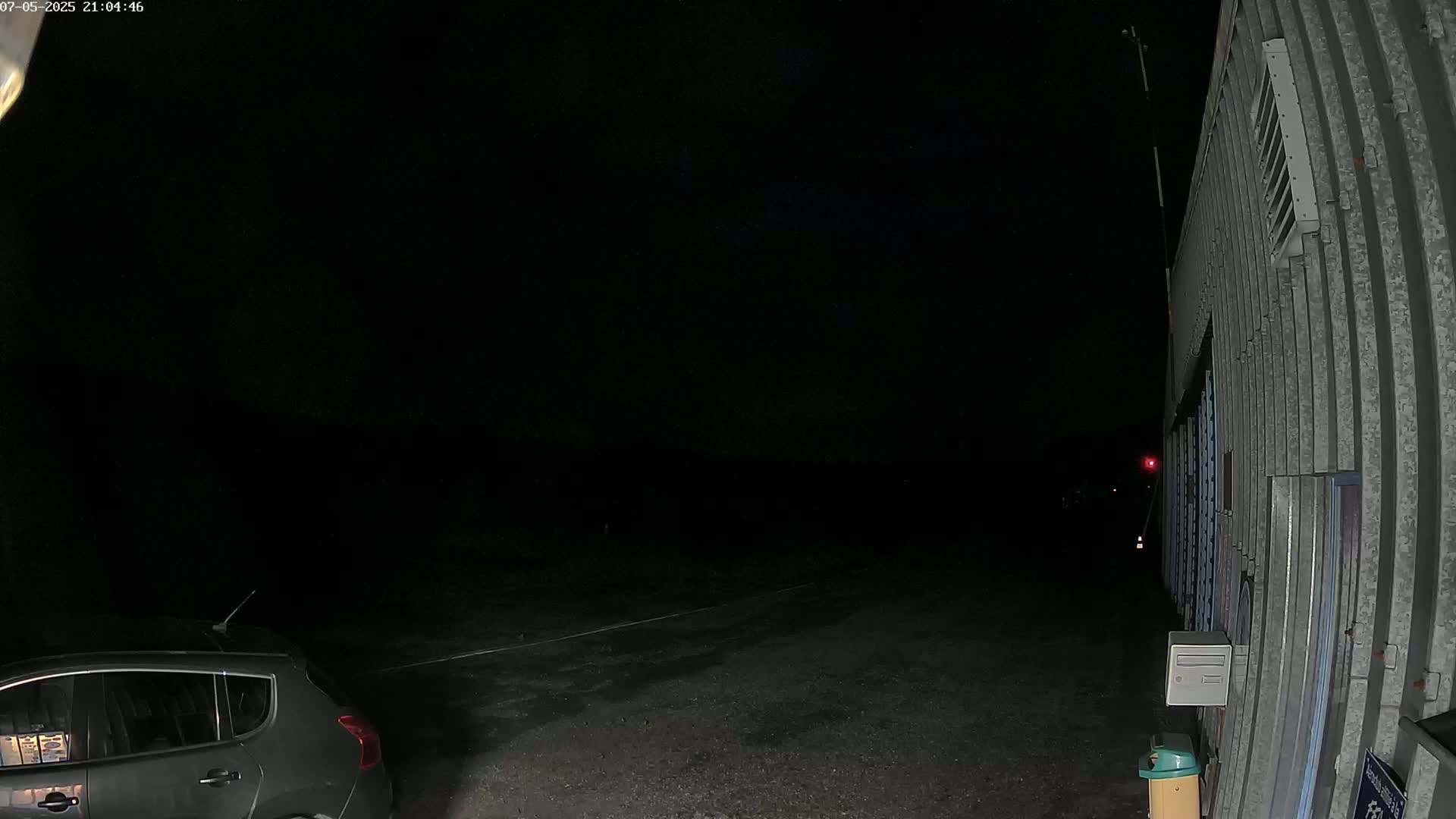 A dark gray car is parked in a dark, empty lot next to a corrugated metal building under a night sky.