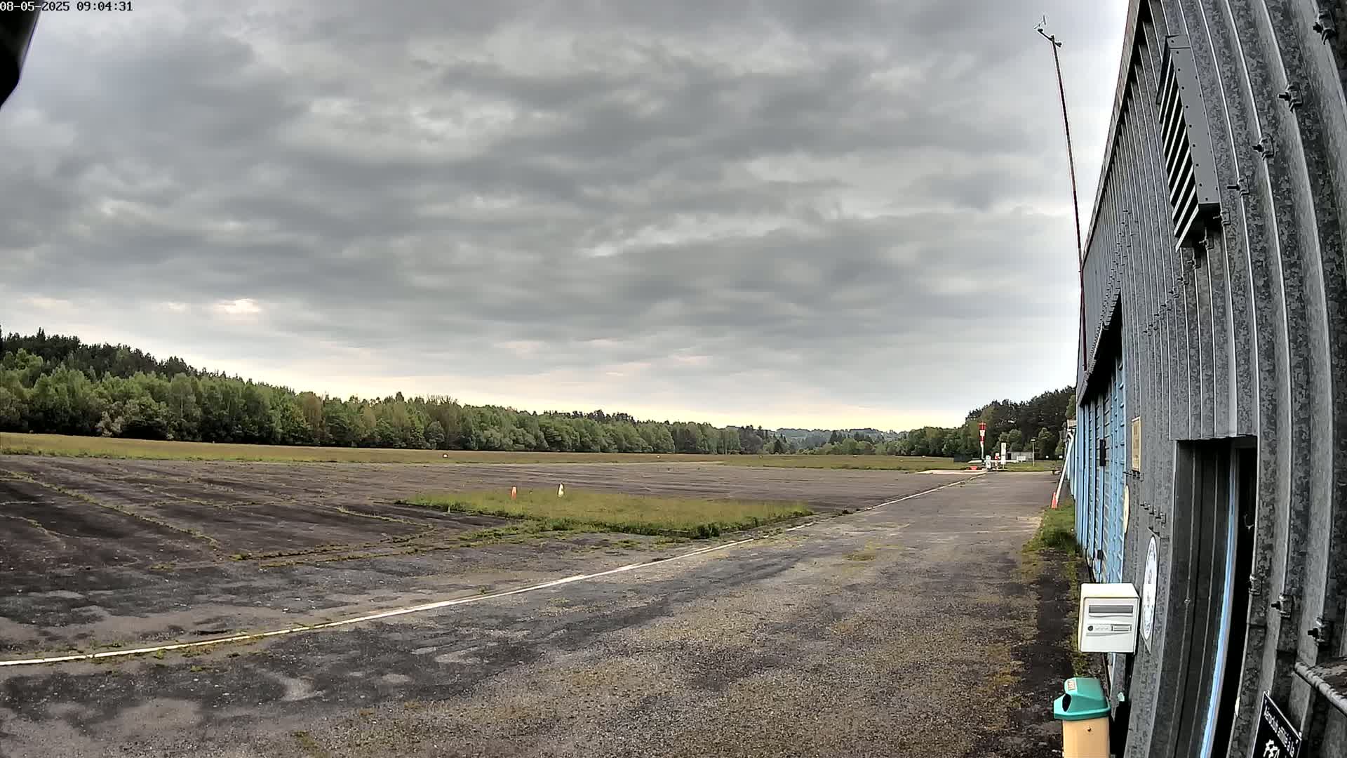 A mostly overcast sky hangs over a long, flat, paved area bordered by a line of trees and a corrugated metal building.