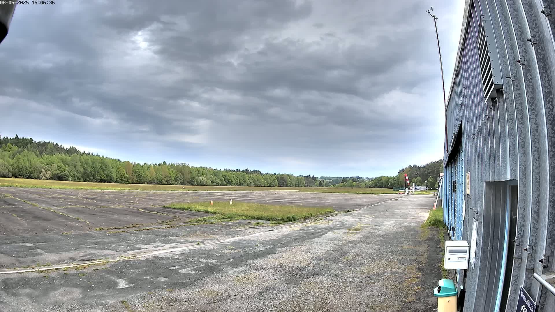 A mostly overcast sky hangs over a flat, cracked tarmac area bordered by trees and a corrugated metal building.