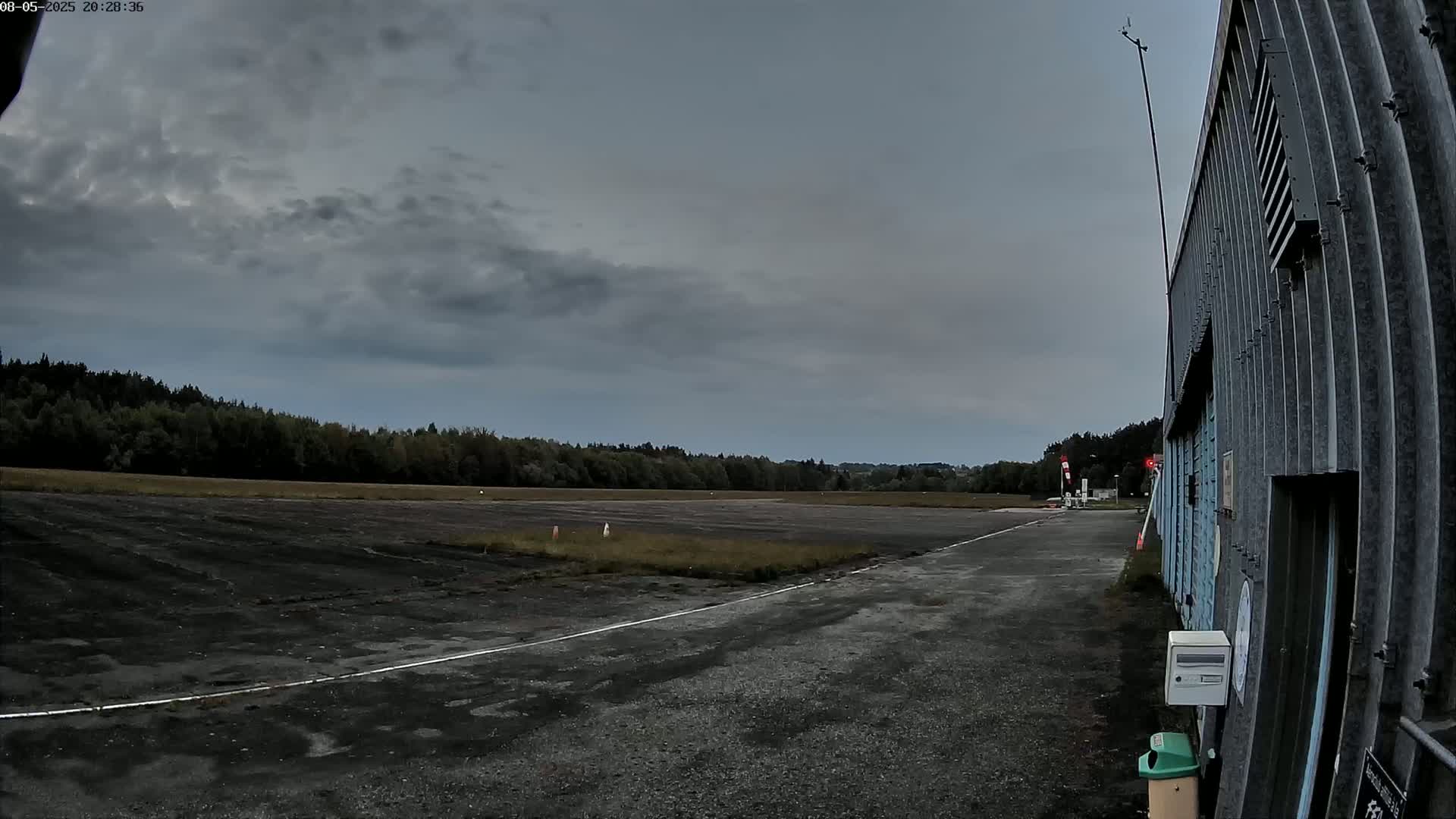 A dark gray overcast sky hangs over a paved airstrip that leads to a corrugated metal building.