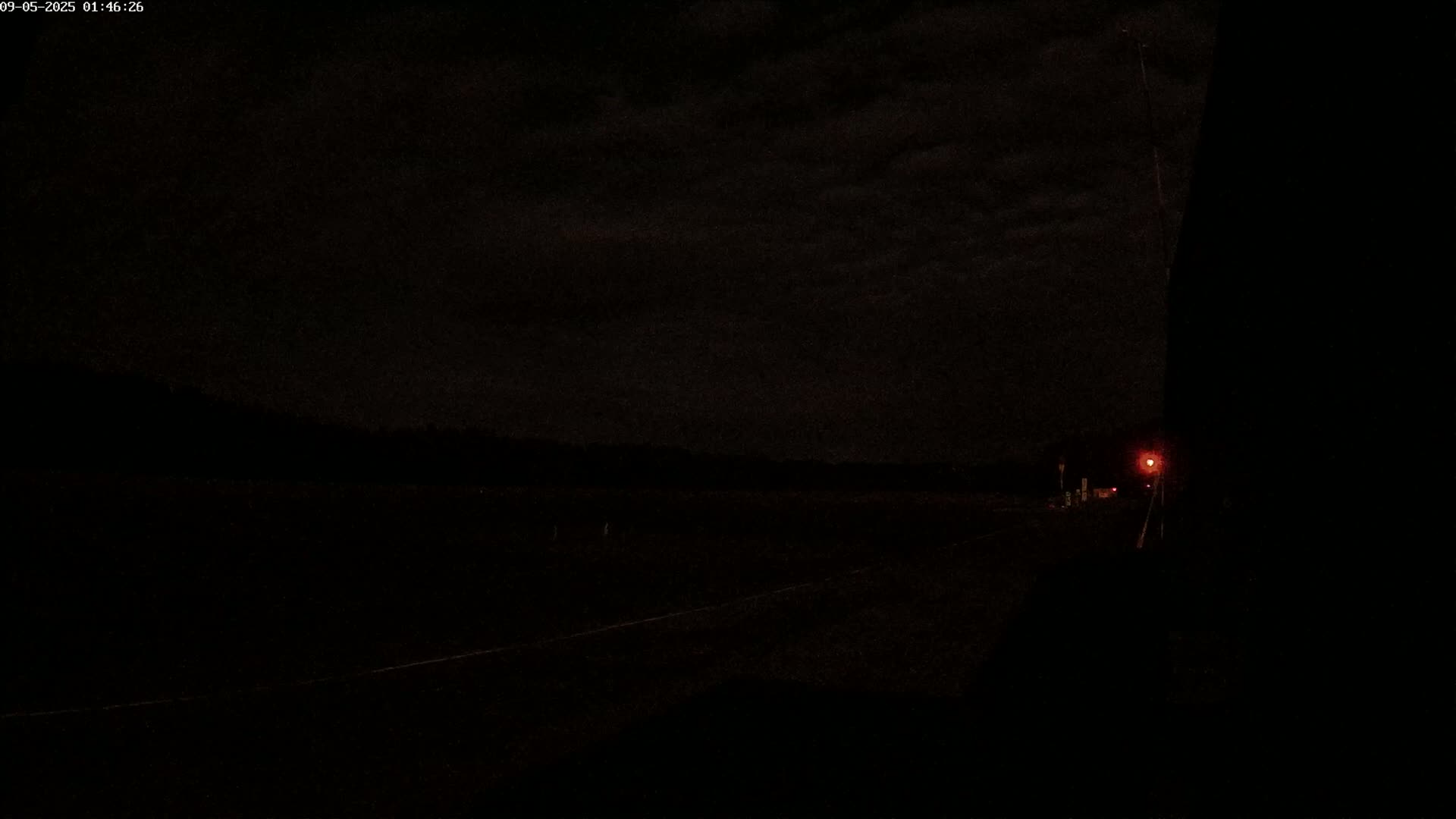 A dark, nighttime road scene shows a faintly visible road with a single, distant red light under a cloudy sky.