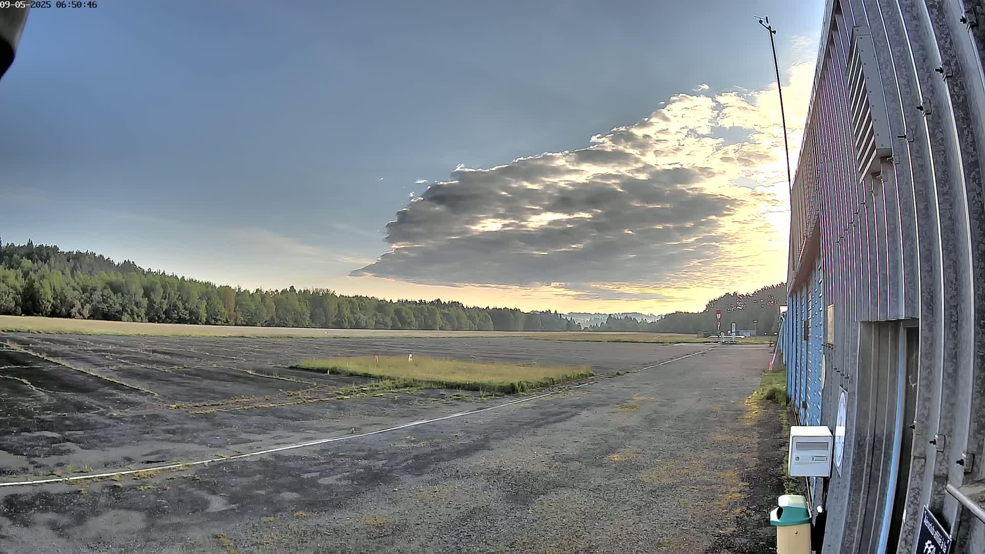 A partly cloudy sunrise illuminates a long, dark runway bordered by a line of trees and a corrugated metal building.