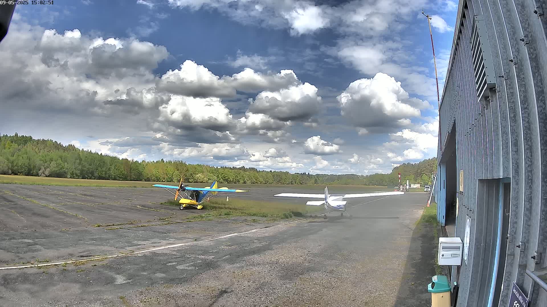 Two small airplanes are parked on a tarmac next to a hangar under a partly cloudy sky.