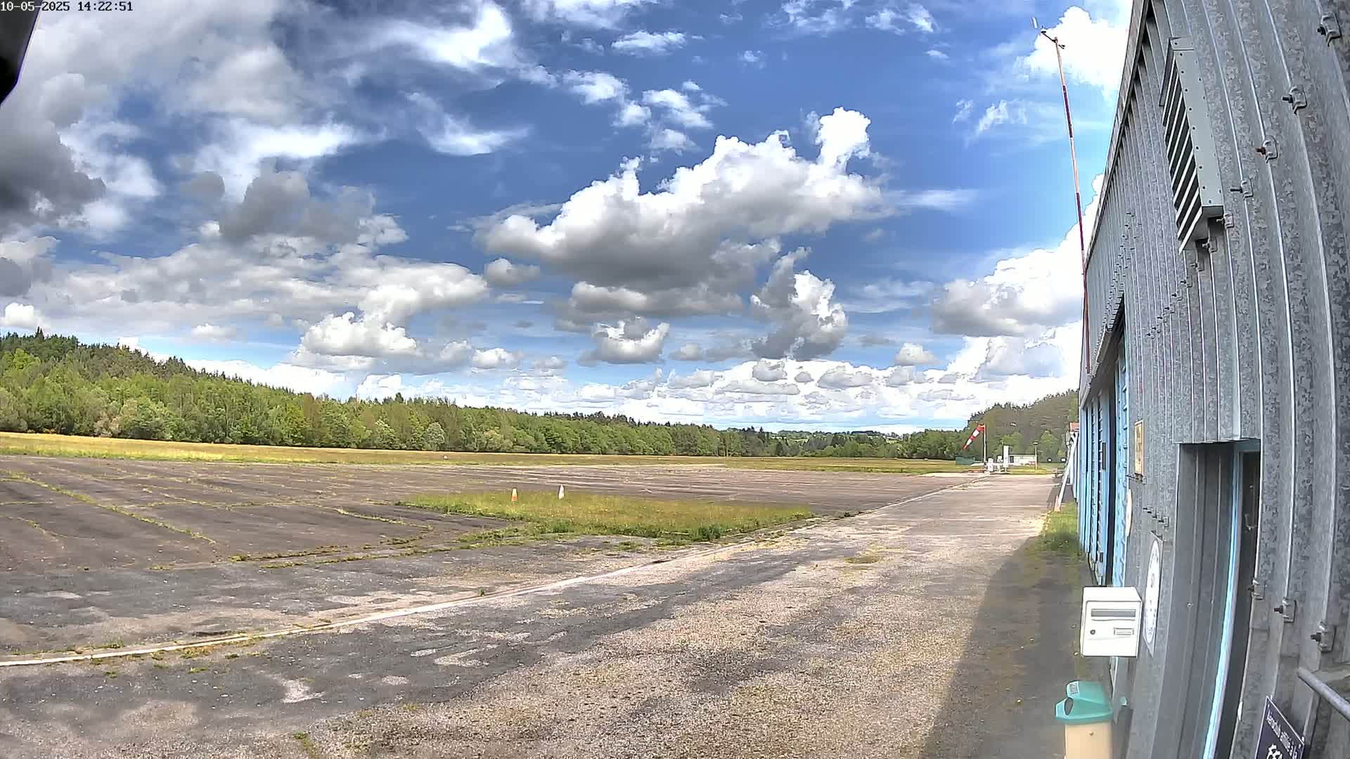 A partly sunny day shows a view of a tarmac area next to a metal building, with trees and a grassy field in the background.