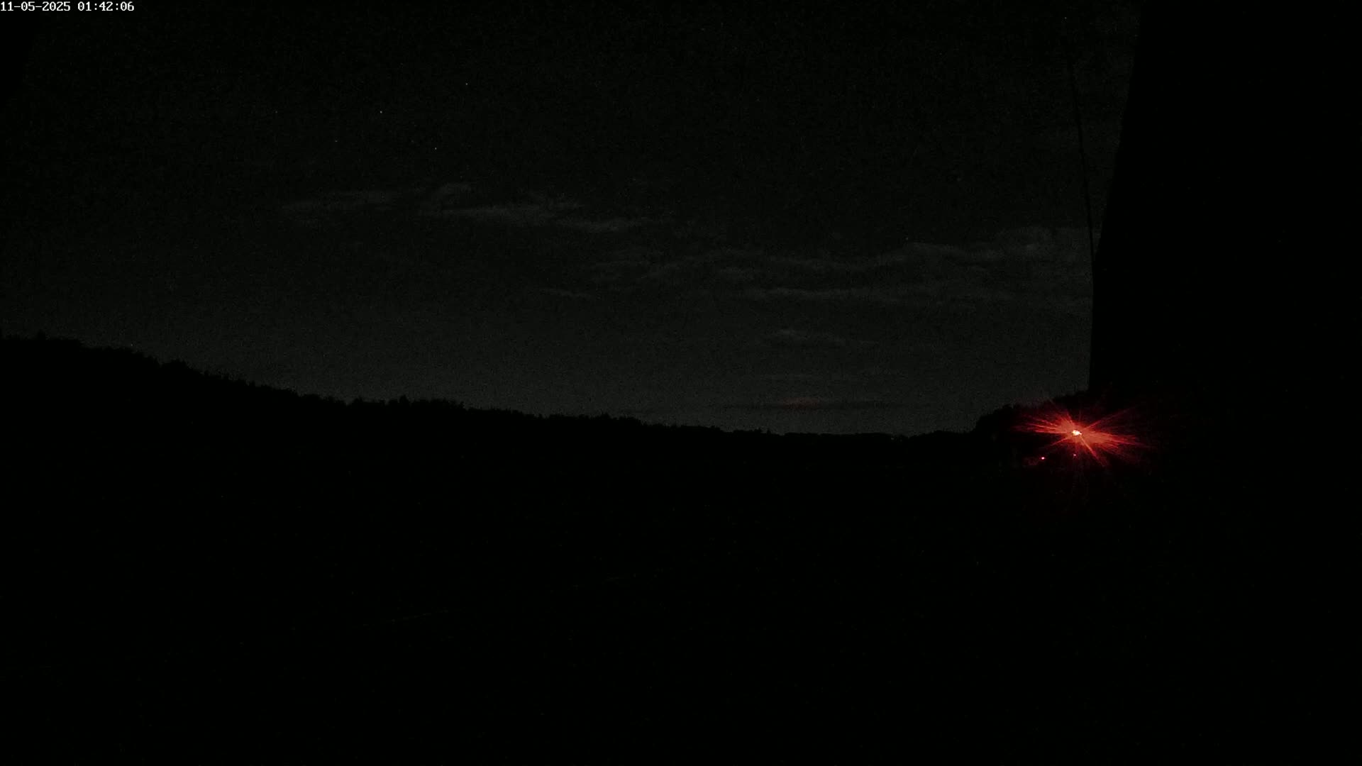 A nighttime landscape shows a dark silhouette of trees and a hill under a slightly cloudy sky, with a bright red light emanating from the lower right.