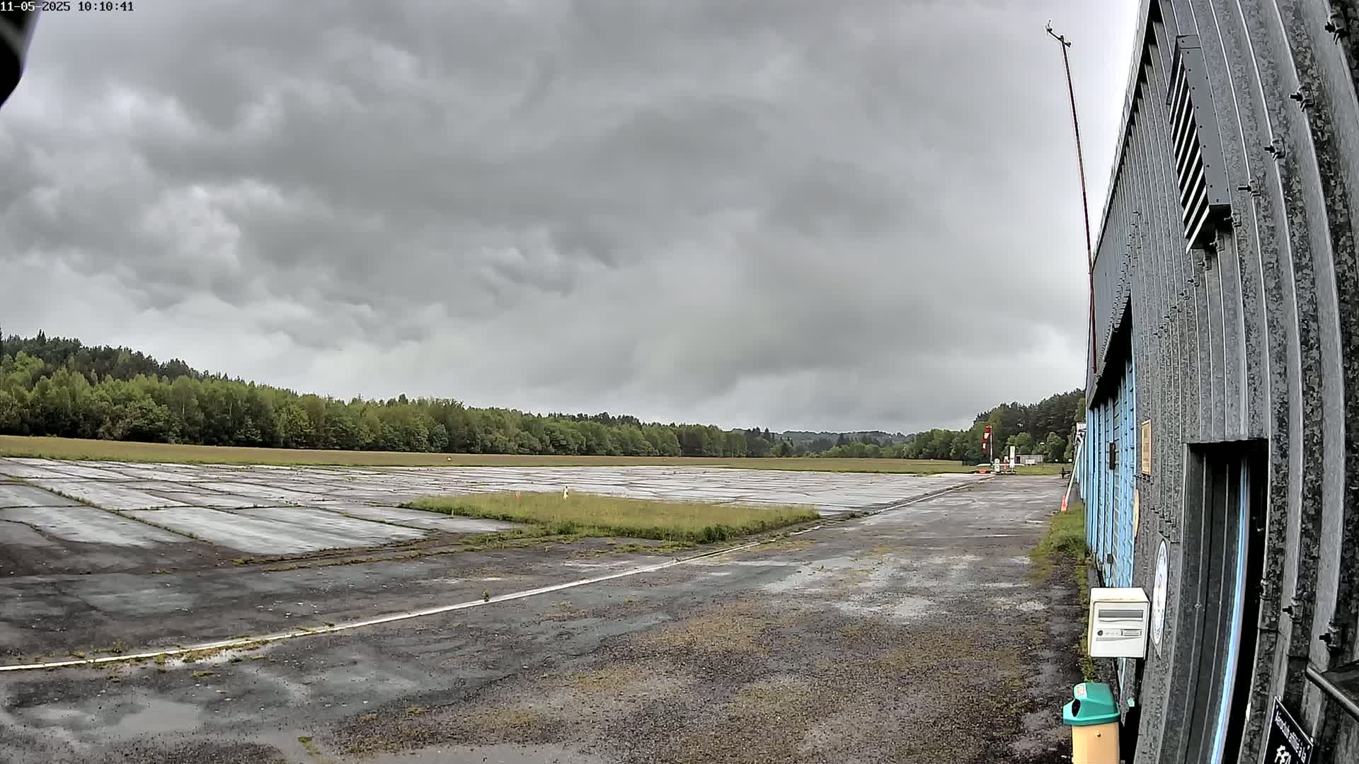 A view of a mostly empty airfield under a cloudy sky shows a long, cracked tarmac surface extending to a line of trees in the distance, with a corrugated metal building to the right.