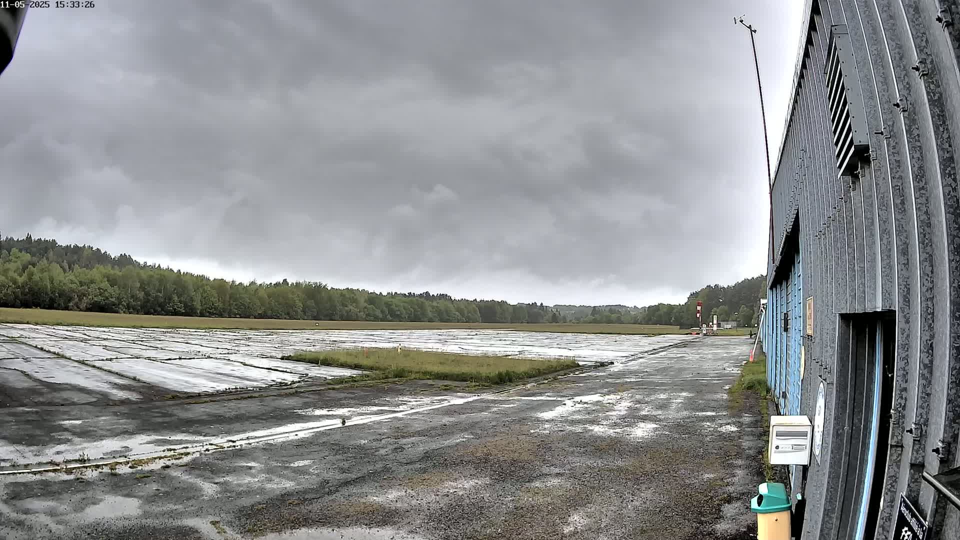 A wet, paved area stretches toward a line of trees under a gray, overcast sky, with a corrugated metal building visible on the right.