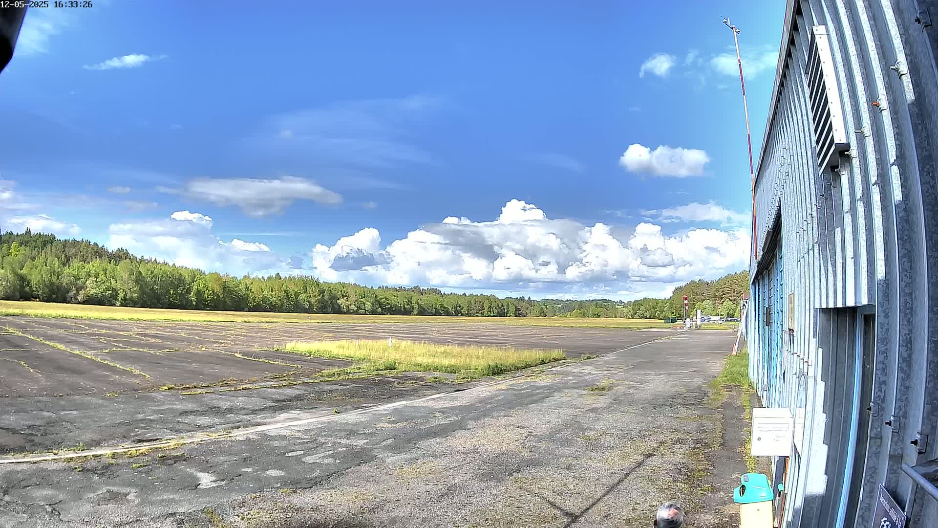 A mostly sunny day with puffy cumulus clouds over a paved airfield bordered by trees and a corrugated metal hangar.