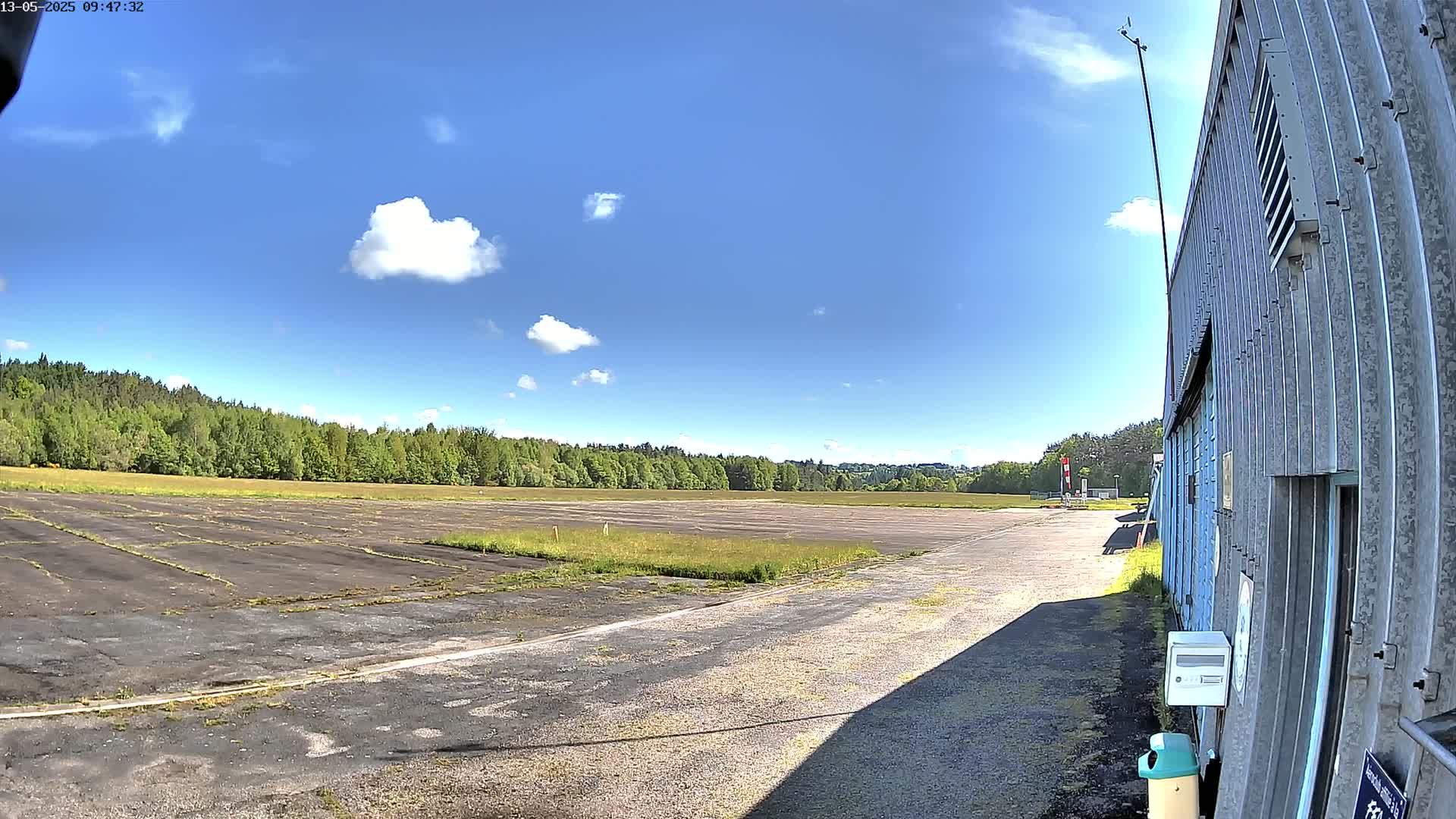 A mostly sunny day reveals a paved, grassy airfield next to a corrugated metal building, with trees lining the horizon.