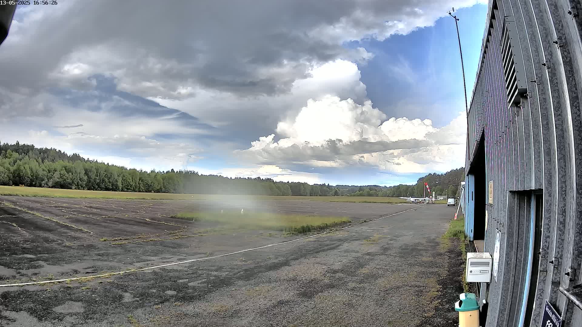 A small airplane sits on a tarmac next to a hangar, with a partly cloudy sky and a line of trees in the background.