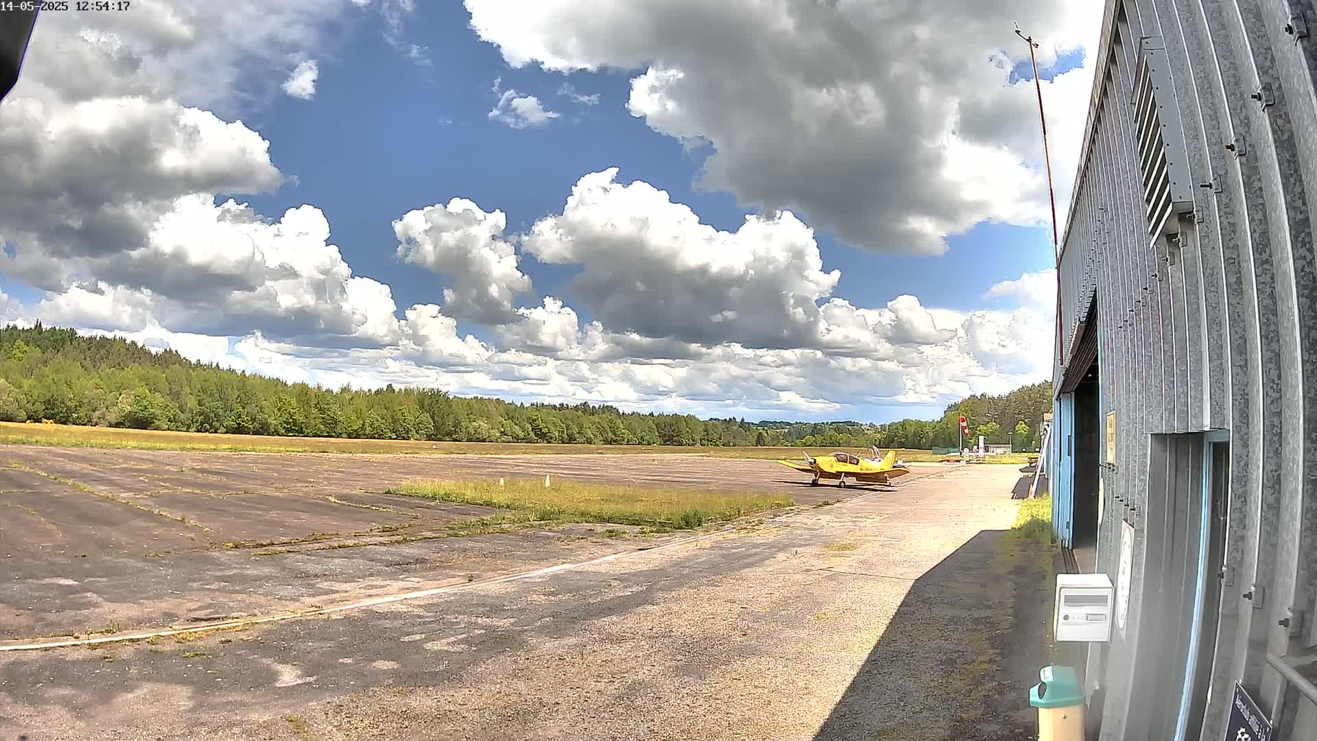 A small yellow airplane sits on a tarmac next to a hangar under a partly cloudy sky.