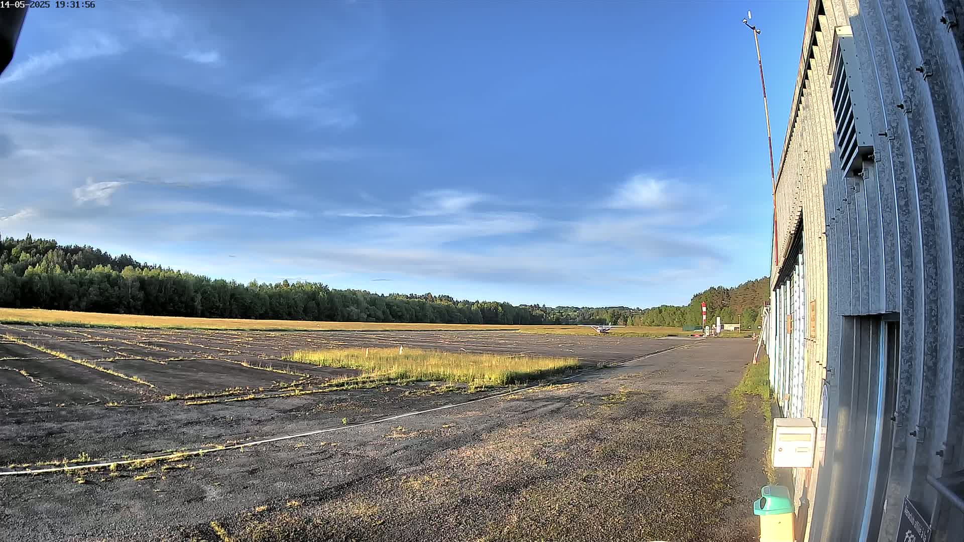 A small airplane sits on a mostly empty airfield under a partly cloudy, sunny sky, next to a weathered hangar.